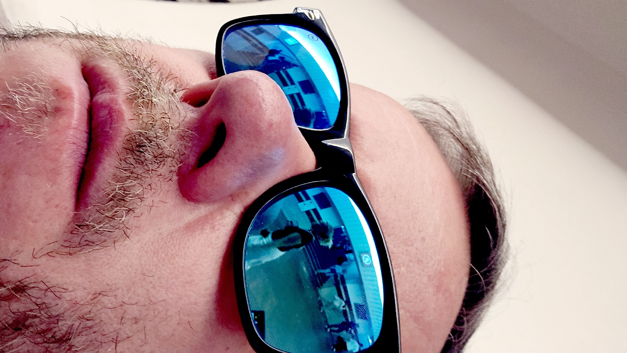A low-angle, close-up shot shows a portion of a person's face, including the nose, lips, and part of the cheek, turned toward the viewer. The person has a light complexion and sparse facial hair. The lips are pink, and the nose is prominent. Black-framed sunglasses with blue mirrored lenses are perched on the nose. The reflection in the lenses suggests an indoor environment, possibly a store or office, with people and monitors visible. The light is generally bright and diffused. The location is not Moscow, Russia, but a retail store with people browsing.