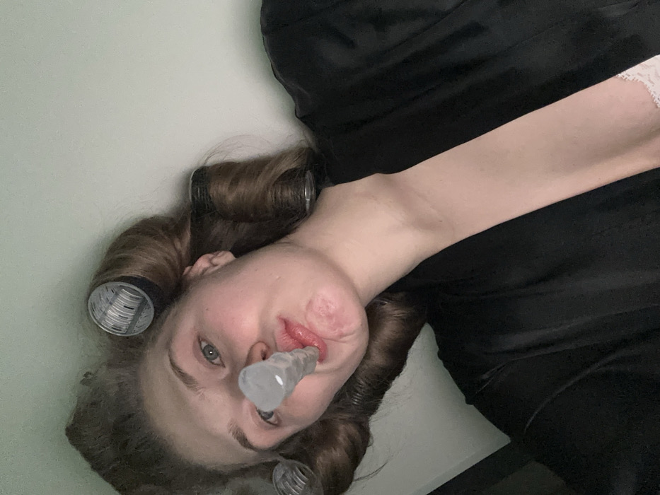 A woman with hair rollers in her brown hair is angled facing down. She is wearing a black outfit, and a clear plastic device is in her mouth, resting against her lips. Her skin is fair, and she has light-colored eyes. The background is a plain light color, possibly a wall.