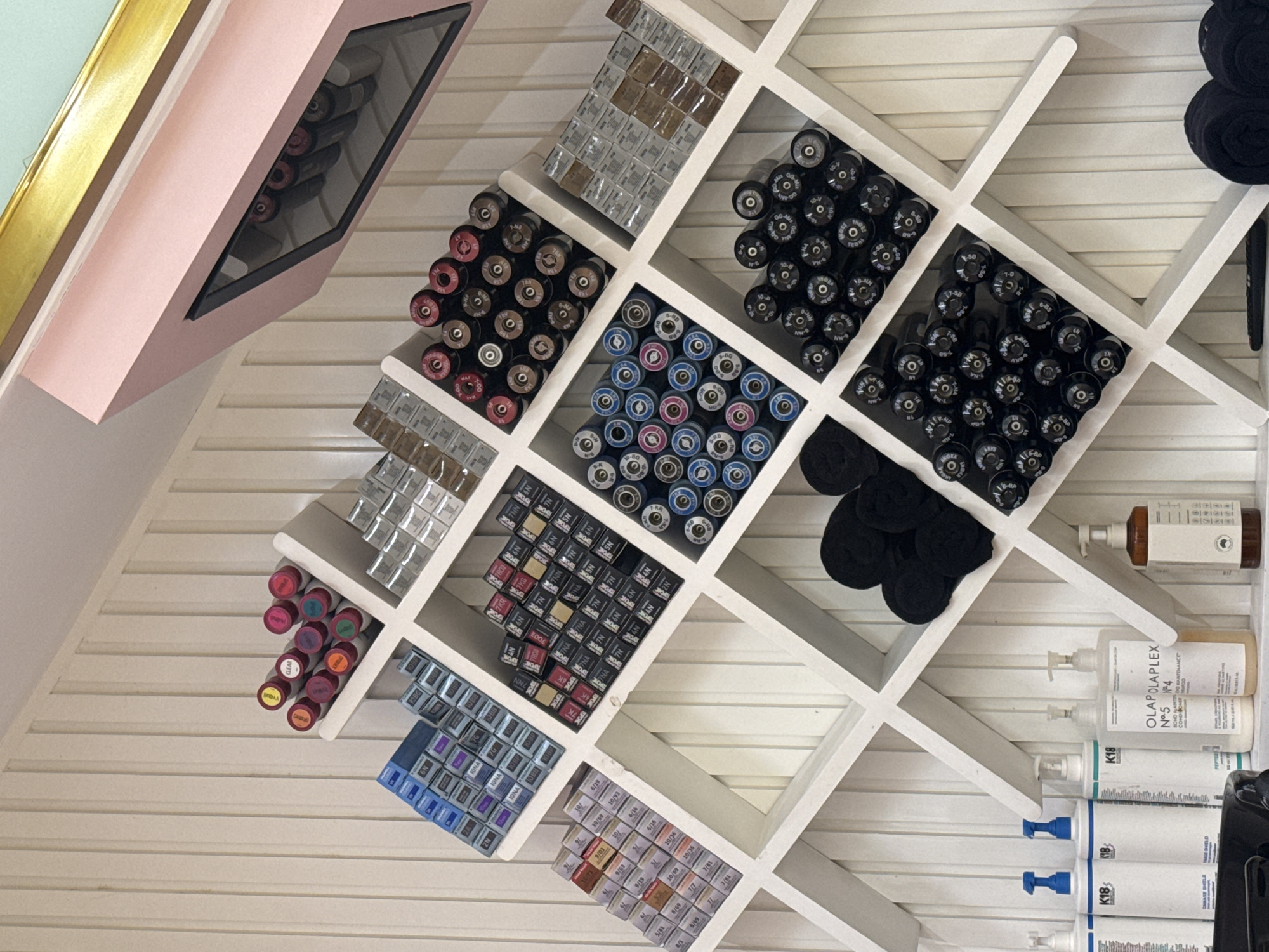 The image shows a slanted, built-in white shelf unit attached to a wall. The unit is filled with hair care products. These products include tubes of hair color in various shades, bottles of Olaplex and K18 products, and black salon hair clips. Above the unit, there is a small display case with more tubes of hair color inside. The setting appears to be a hair salon, though the specific location in North Kuta, Indonesia, cannot be determined from the image alone.
