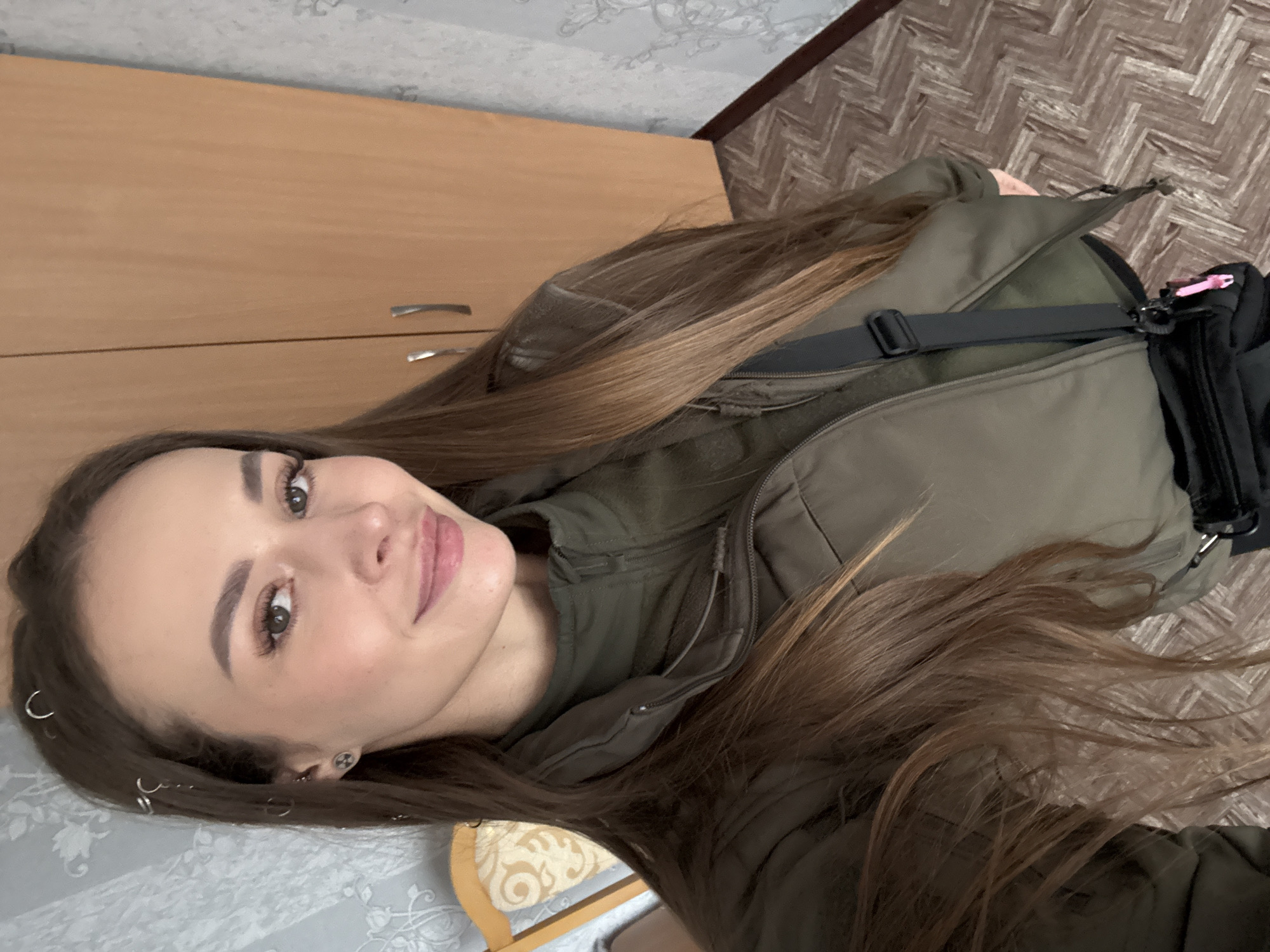 A woman is posing indoors in an off-center, rotated selfie. She has long brown hair, fair skin, and visible makeup. She is wearing a green, quilted jacket with a black strap running across her chest. A black bag with a pink detail is visible near the jacket.
The background includes a light-colored wooden cabinet, patterned wallpaper, and a herringbone-patterned floor.
The location is not evident in the image, and there is no indication it is Chornobyl, Ukraine.