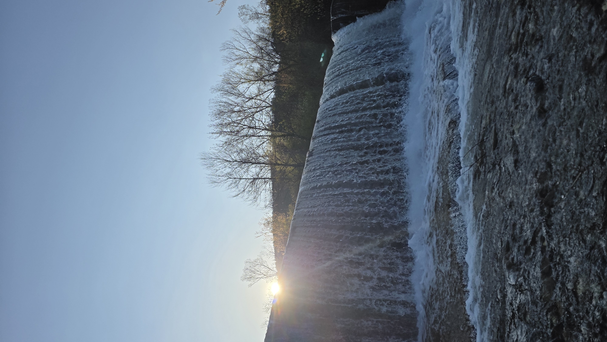 The image depicts a waterfall or dam structure with water cascading down. To the left, there are trees with bare branches against a clear blue sky. The sun is visible behind the structure, creating a lens flare effect. The location is not specified in the image but it could be in the city of Blagoevgrad, Bulgaria.