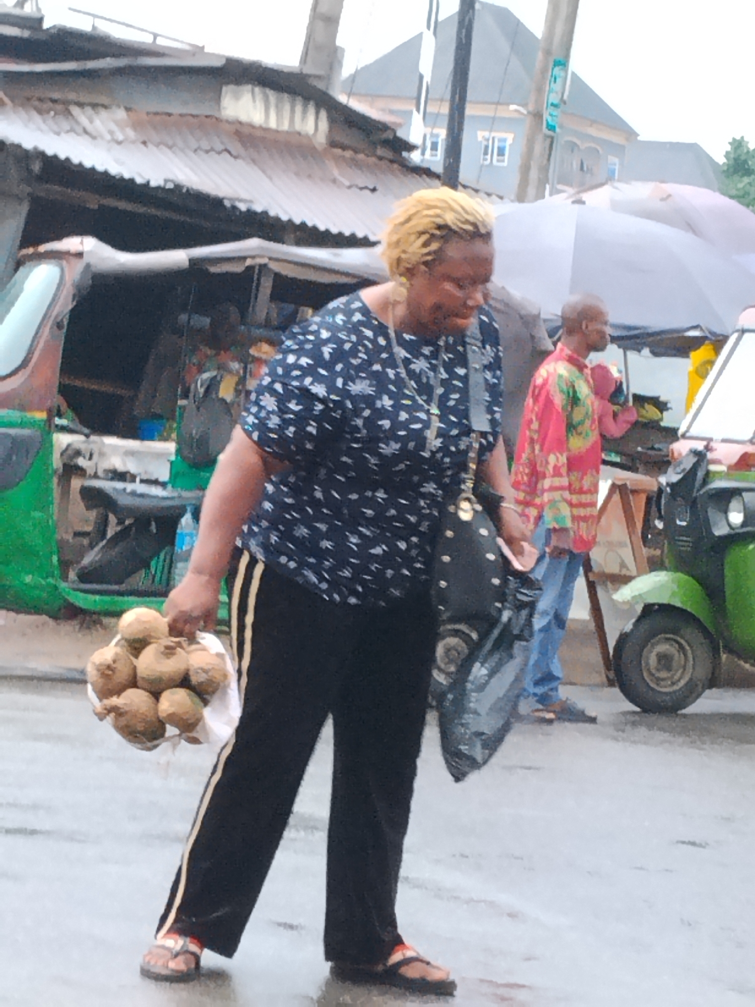 In the city of Aba, Nigeria, an African woman with blonde hair is walking. She is wearing a navy blue floral print shirt, black pants with gold stripes, and sandals. She is carrying a bag of brown fruits, possibly coconuts, in her left hand and a black purse on her right shoulder. Behind her, a man in a red and green patterned shirt and blue jeans is standing. Two green "keke" tricycles, commonly used as taxis in Nigeria, are visible in the background. The street appears wet, suggesting recent rain. A shop with a corrugated iron roof is to the left, and a building is in the distance.