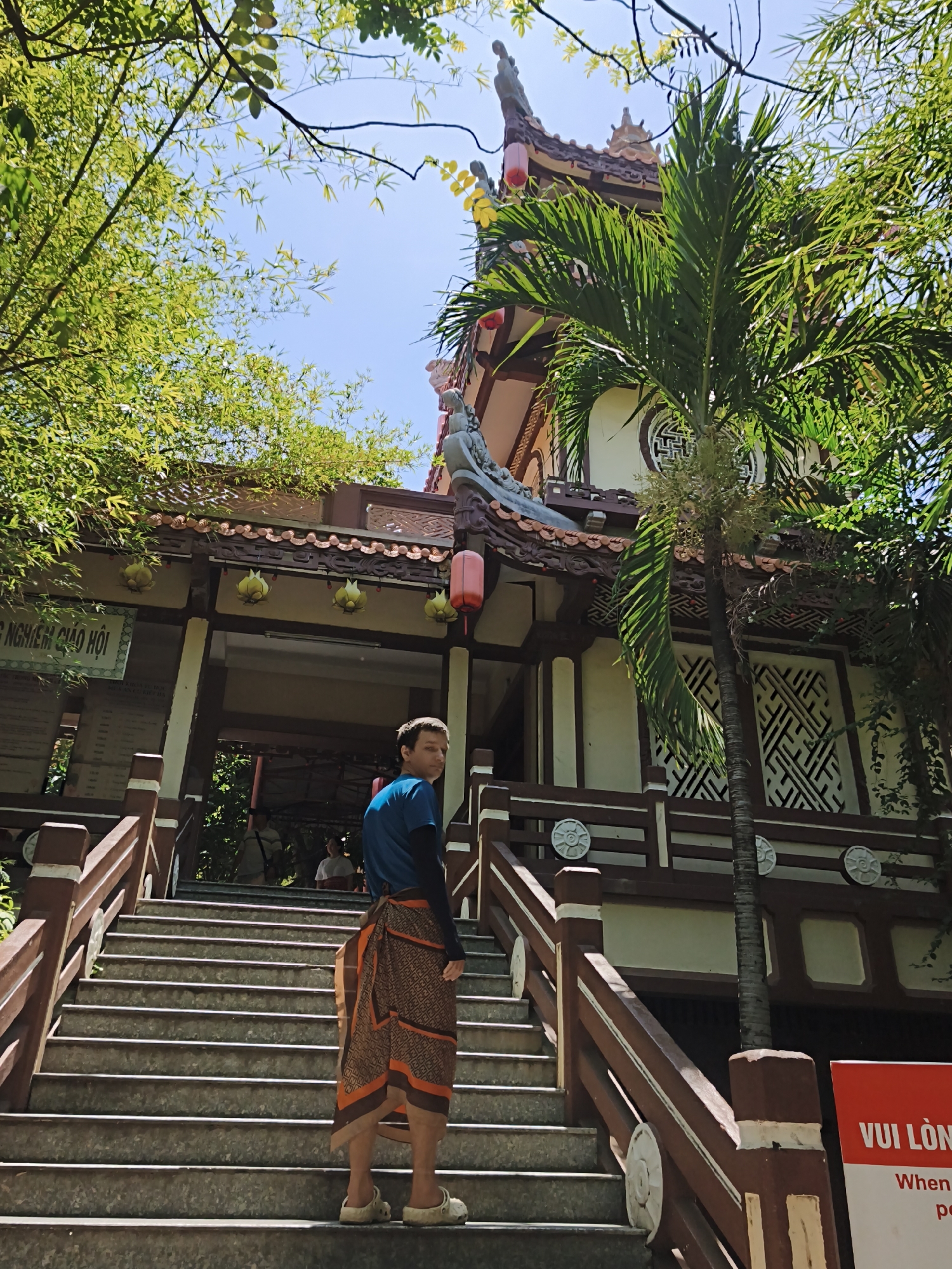 A person stands on stone steps leading up to a building in Tay Nha Trang Ward, Vietnam. The person is viewed from behind and is wearing a blue t-shirt, black arm sleeves, and a brown and orange patterned sarong. They are also wearing light-colored clogs. The building appears to be a temple or pagoda, with ornate architecture including a tiled roof, decorative eaves, and lanterns. Lush green trees and bamboo surround the building, with a palm tree in the foreground. A red sign with white text is visible on the right side of the steps. The sky is clear and blue.