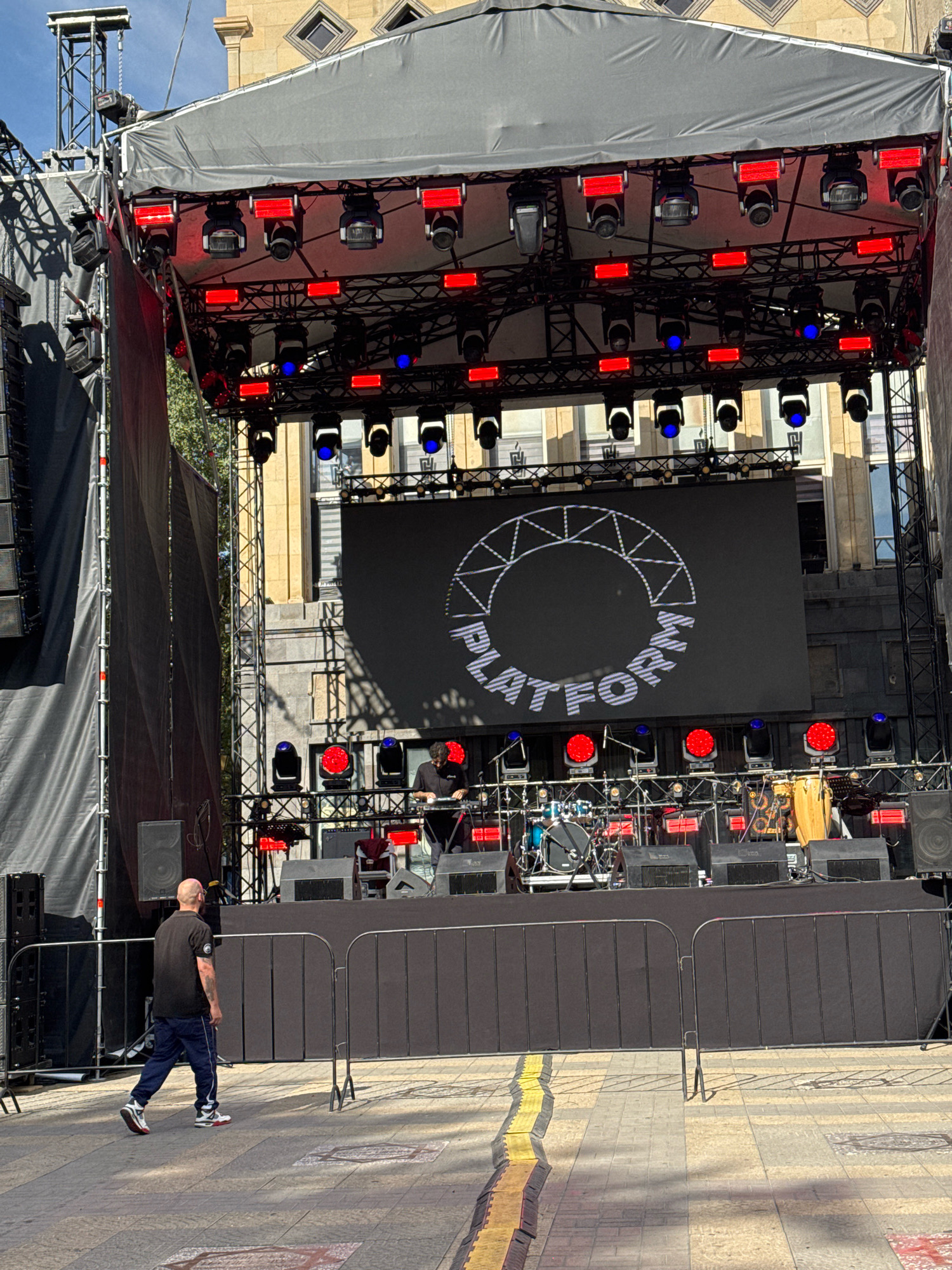 An outdoor stage setup is shown in front of a building, likely in the city of Yerevan, Armenia, due to its architectural style. The stage is covered by a black tarp and equipped with multiple rows of lighting fixtures, including red and blue lights, and a large central LED screen displaying a circular graphic with the word "PLATFORM" integrated within it. To the left of the stage, large black speaker systems are visible, along with tall metal scaffolding and black fabric. On the stage itself, a drum set and a conga drum are set up, along with musical equipment and speakers. Two individuals are present: one person, a bald man wearing a black t-shirt and blue pants, is walking away from the stage towards the viewer, and another person, partially obscured, is seen operating a keyboard on the stage. A temporary metal barrier runs across the foreground, separating the stage area from the viewer's perspective. The ground is paved with stone tiles, and the overall impression is of a pre-performance or setup phase for an outdoor event.
