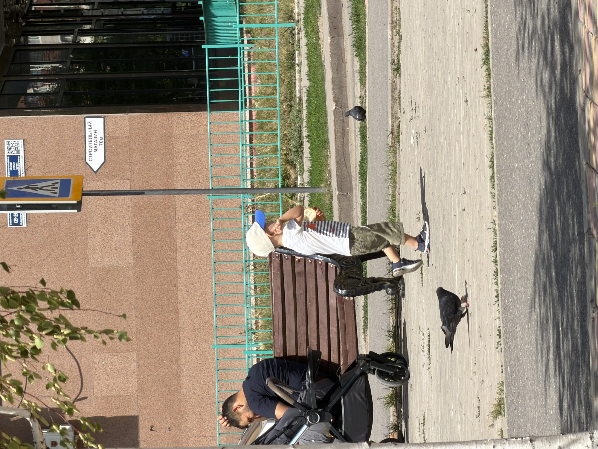 In the city of Astana, Kazakhstan, on a sunlit sidewalk, a young boy wearing a white t-shirt, khaki shorts, a white baseball cap with a blue brim, and dark sneakers, stands facing right, holding a piece of bread in his right hand and touching his face with his left. He is walking on a light-colored paved surface with scattered debris. To his left, a man in a dark t-shirt and shorts sits on a wooden bench, leaning his head against his hand, next to a dark-colored stroller. Behind them, a teal metal fence runs along the edge of a grassy area. A dark gray pigeon is walking on the sidewalk in the foreground, and another pigeon is visible further in the background near the fence. A building with a textured brown facade and large windows forms the backdrop. A street sign pointing to the right with Russian text that reads "СТРОИТЕЛЬНЫЙ МАГАЗИН 70М" (Construction Store 70M) is mounted on a pole to the left of the building. A white tree trunk is partially visible in the left foreground.