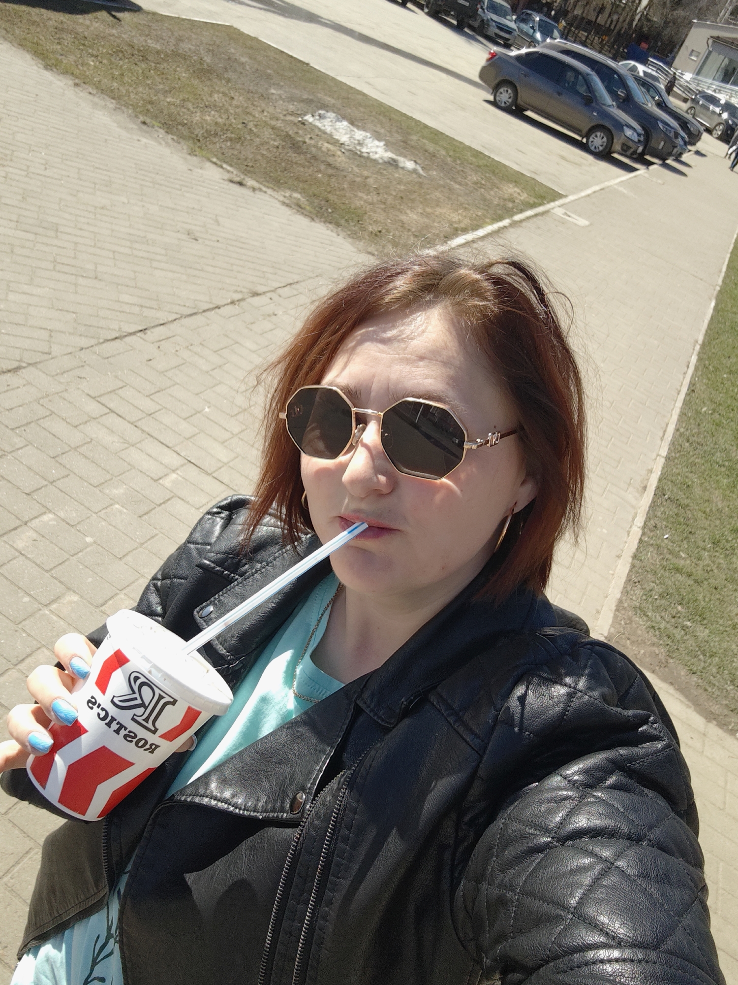 A woman takes a selfie while drinking from a paper cup with a straw on a sunny day in Chaikovsky, Russia. She is wearing a black quilted leather jacket, sunglasses, and a light blue top. The cup has red and white stripes with a logo that reads "KOCTOP". The background shows a sidewalk made of grey paving stones, a patch of dead grass, a street, and several parked cars. The overall scene suggests it is springtime.