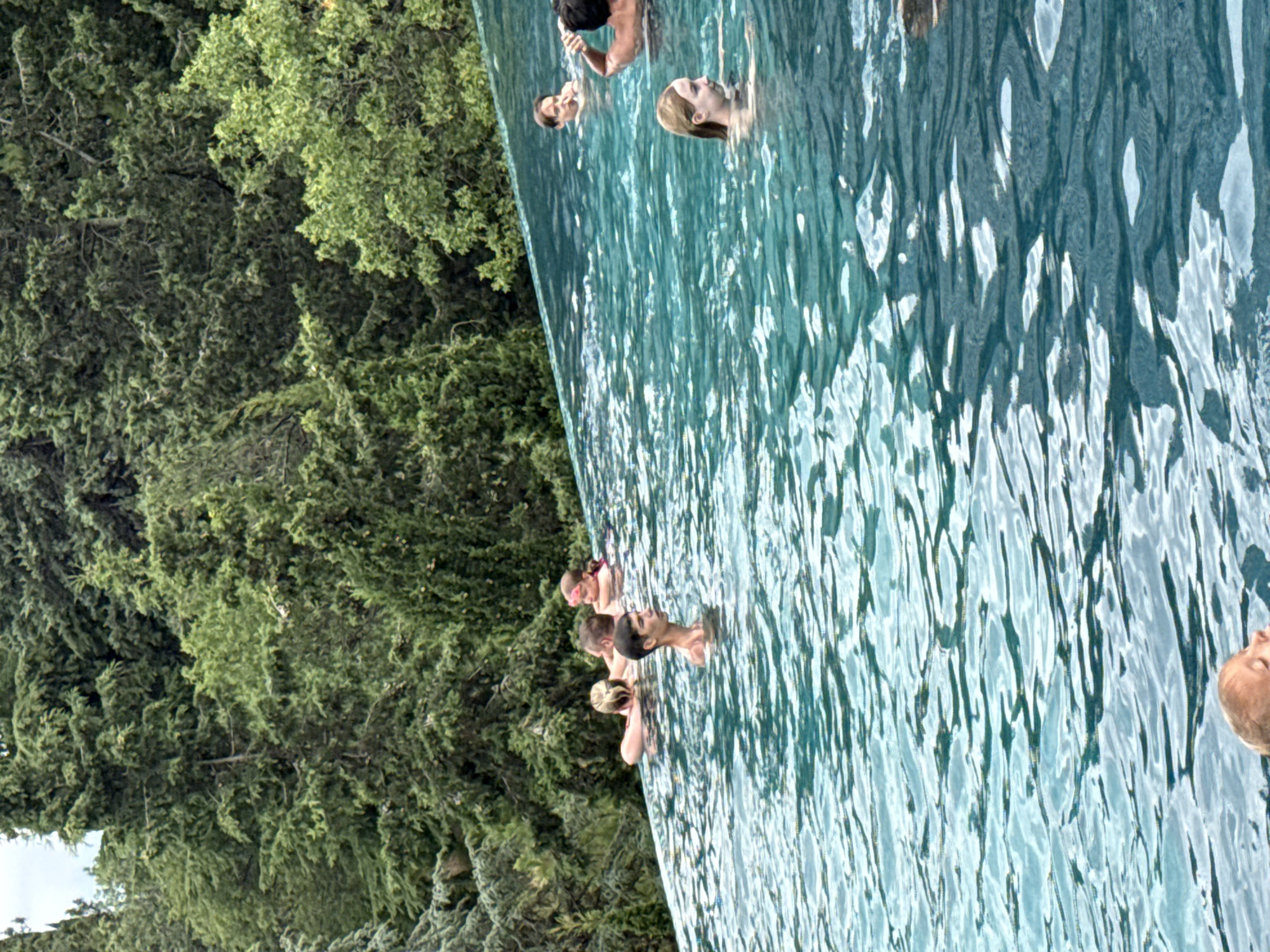 An outdoor infinity pool with a backdrop of lush green trees. Several people are visible in the pool, including adults and children. The water is a vibrant turquoise color and reflects the surrounding greenery and sky. One person, a young child, is in the foreground, with their face mostly submerged in the water. In the middle ground, a young man stands in the water, looking to the side. Further back, a group of people are swimming and interacting, including a woman with blonde hair and goggles on her forehead. The pool appears to be a part of a resort or private residence, set in a natural, wooded environment. There is no visible signage or indication of the specific location in the city of Yalta, Ukraine.