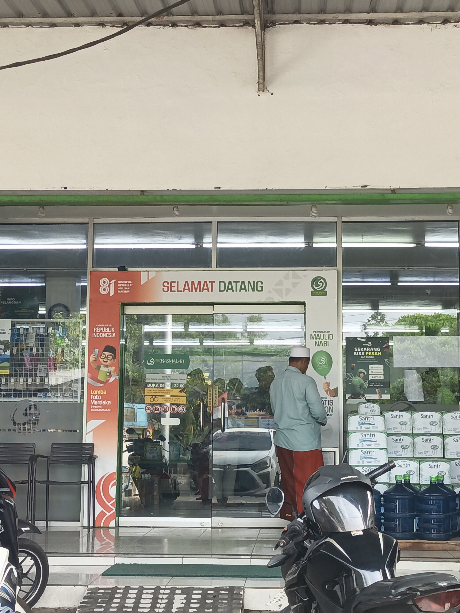 The image depicts the entrance of a convenience store in Baujeng, Indonesia. A man wearing a light blue long-sleeved shirt, red pants, and a white skullcap is standing inside the store, facing away from the camera. He appears to be looking at something within the store. In front of the store, several motorcycles are parked. Inside the store, a display of bottled water and packaged goods is visible. The store's signage includes the words "SELAMAT DATANG" (Welcome) and "BASMALAH". The overall environment suggests a typical commercial street scene in Indonesia.
