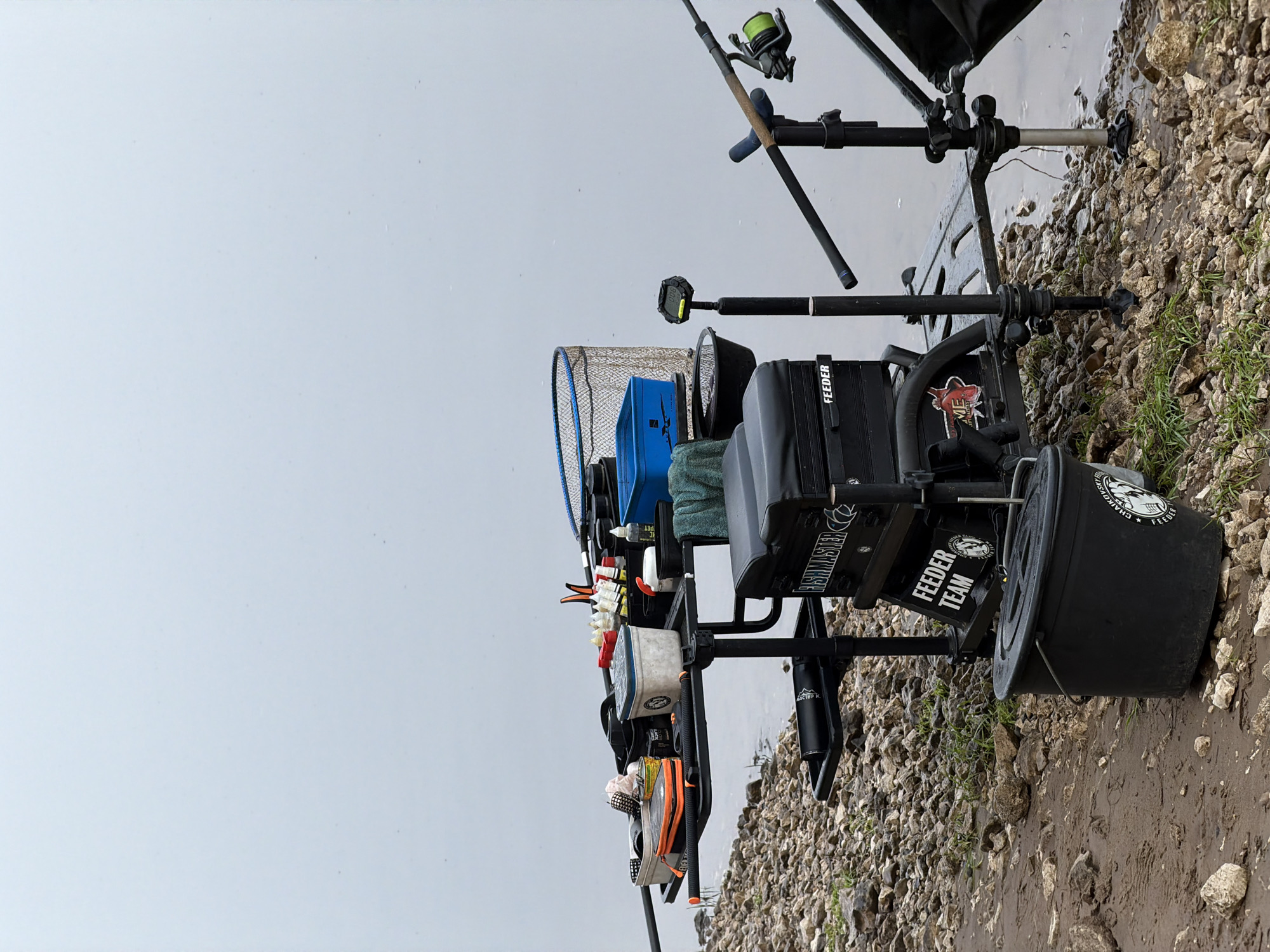 The image displays a comprehensive fishing setup, rotated 90 degrees counter-clockwise. When viewed upright, a fishing station is positioned on a rocky and muddy bank, adjacent to a calm body of water under an overcast sky, in Noviy, Russia.
The scene focuses on an elaborate fishing rig, implying an active or prepared feeder fishing session. No individuals are visible. Dominant subjects include multiple fishing rods equipped with reels, positioned in rod rests. A large blue-rimmed landing net is prominently placed, alongside a multi-functional black seat box or tackle station, which has various attachments and compartments. Several black buckets, smaller containers, and diverse tackle items are arranged around the station.
The weather appears overcast, with a uniformly light gray sky reflecting on the smooth surface of the water, indicating calm conditions. The bank consists of a mix of small to medium-sized rocks, wet mud, and sparse patches of grass.
Visible text on the equipment includes "FEEDER TEAM", "FEEDER" (on multiple items), "FISHMASTER", and "EXTREME". One large black bucket clearly displays "CHAIKOVSKIY FEEDER" along with a fish logo.