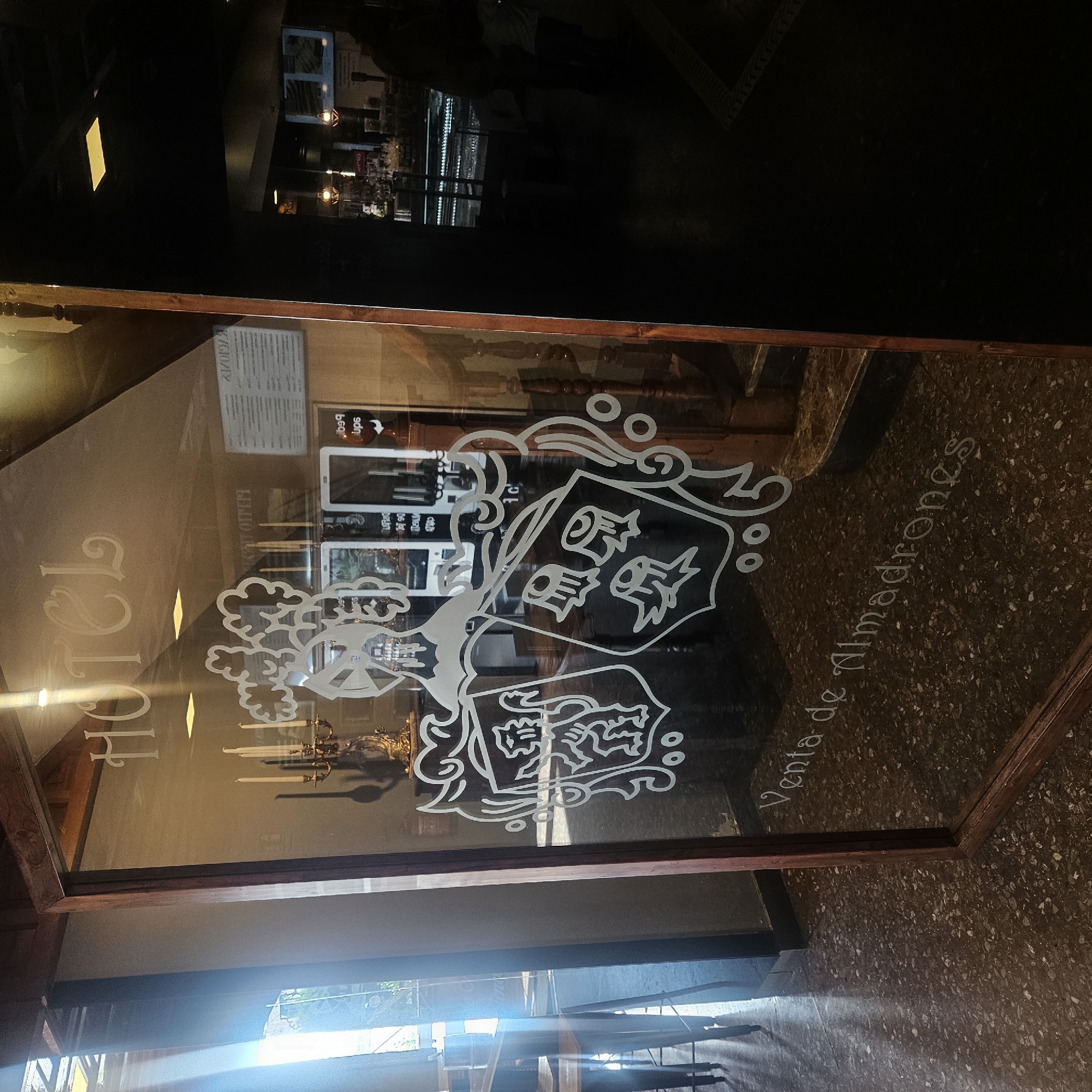 The image shows the entrance to a hotel in Almadrones, Spain. The hotel's name, "HOTEL", is visible on the glass door in a decorative font. Below the name, a coat of arms is etched onto the glass. The text "Venta de Almadrónes" is also etched on the glass, indicating that this is a sales point for products from Almadrones. Inside the hotel, a staircase and a bar area are visible. The flooring in the entrance is a speckled pattern. The lighting in the image is dim, with some natural light entering from the left side.