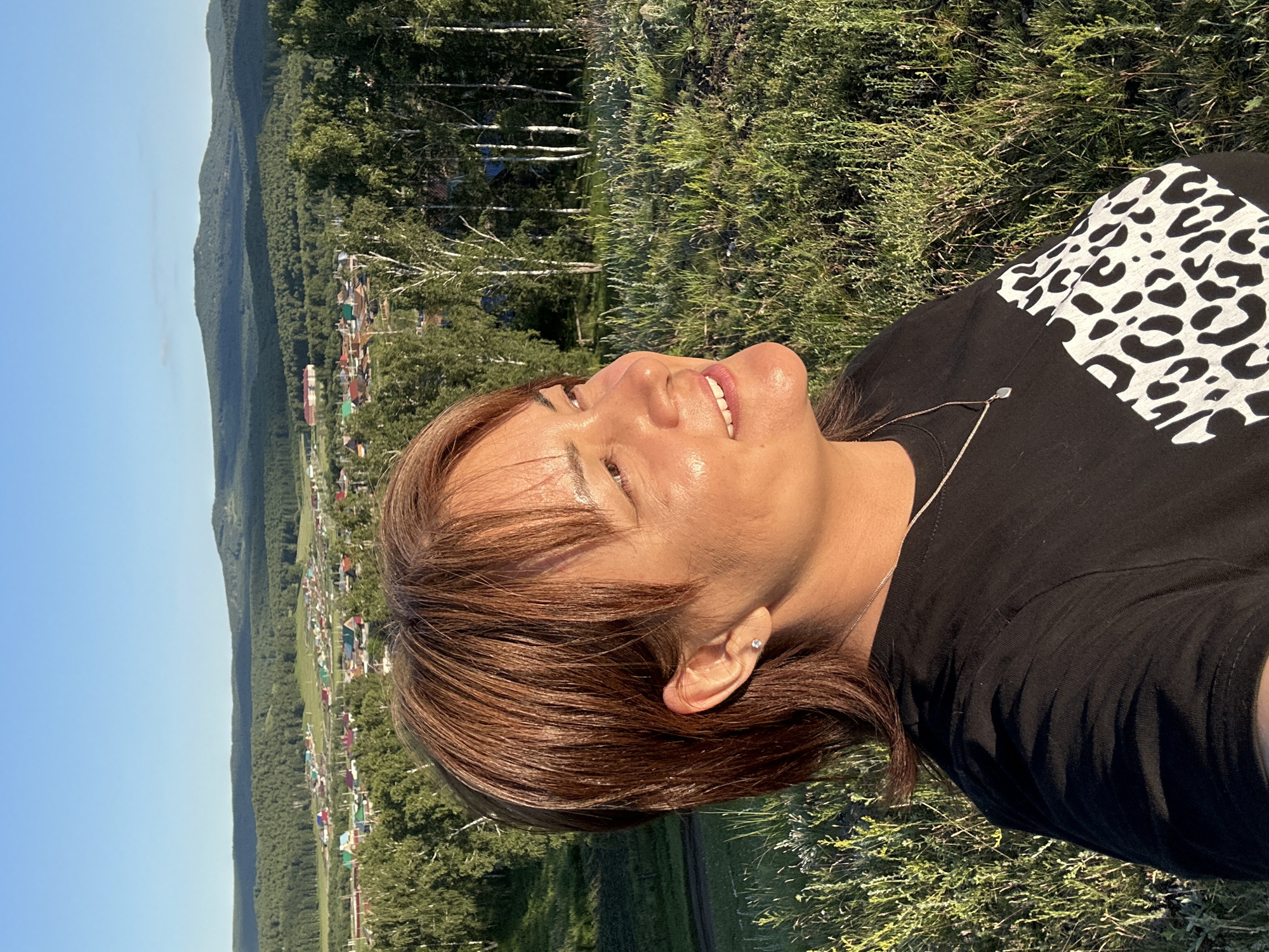 This is a portrait of a person lying down in a grassy area, with a village and forested hills in the background. The person, who appears to be female, has brown hair and is looking upwards and to the side with a slight smile. She is wearing a black t-shirt with a white leopard print design on the chest and a delicate necklace. A small stud earring is visible in her ear. The sun appears to be shining brightly, casting a warm glow on her face. The background shows a scattering of houses with colorful roofs, nestled amongst dense green trees, with rolling hills receding into the distance. The sky is clear and blue. The scene suggests a relaxed and peaceful moment outdoors, possibly during the daytime. No visible text is present in the image.