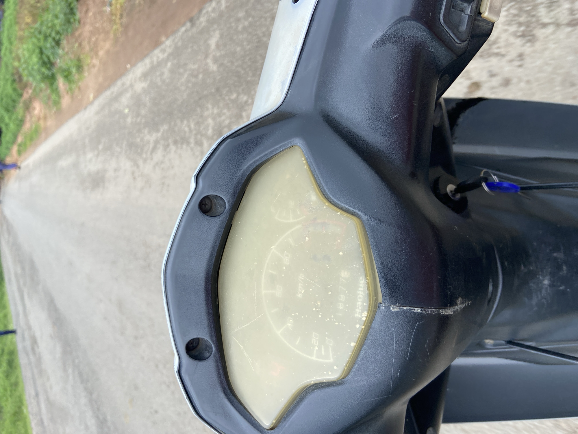 The image shows a close-up of a motorcycle's dashboard, with a blurred road and greenery in the background. The dashboard is black and features a yellowed, scratched display with some markings that appear to be a speedometer and an odometer reading "199776". A key with a blue fob is inserted into an ignition switch. The road ahead is paved and appears to be in a rural or suburban area. The overall impression is that the motorcycle is either parked or moving slowly on a road in Mararaba, Nigeria.