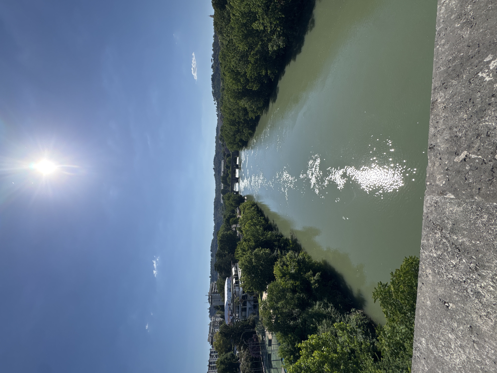 The image shows a view of the Tiber River in Rome, Italy, from a bridge. The river is wide and green, reflecting sunlight. Trees line both banks of the river. On the left bank, buildings are visible. The sky is blue with some clouds. The sun is bright, creating a lens flare effect. Part of a stone bridge structure is visible in the foreground.