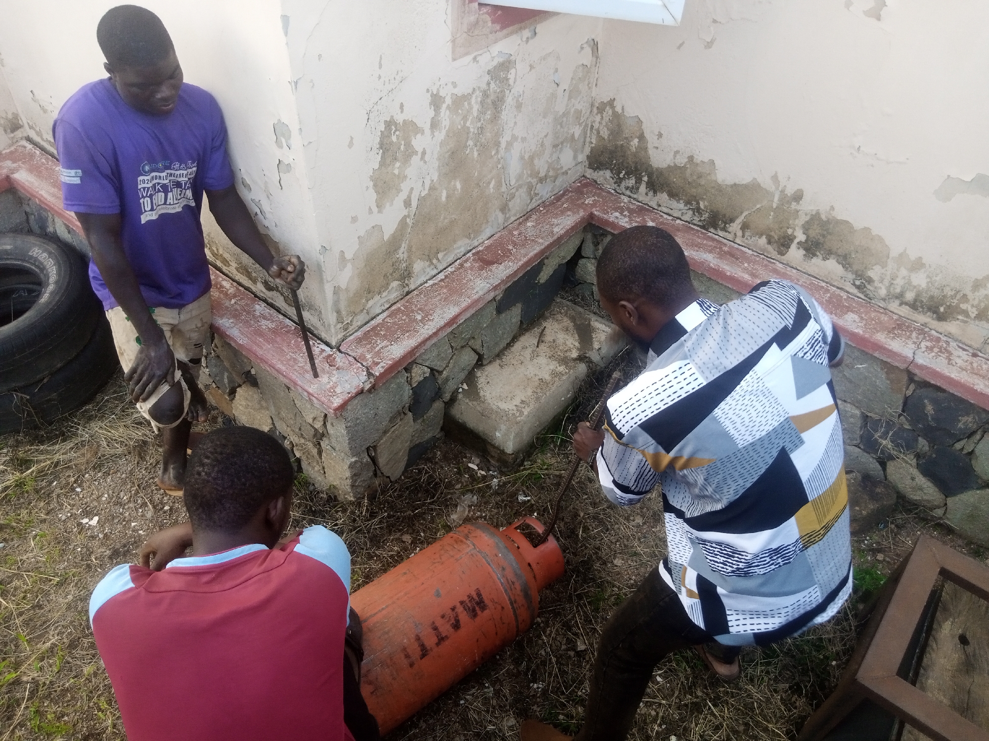 In Jos, Nigeria, three individuals are engaged in an activity near a building with peeling paint. The scene depicts an outdoor setting with dry grass and dirt. One person, wearing a purple t-shirt and light-colored shorts, is using a metal rod to pry at something on the ground. Another person, in a red and light blue shirt, is crouched down, seemingly observing or assisting. The third individual, dressed in a patterned shirt and dark pants, is also holding a metal rod and appears to be working on a large, orange gas cylinder lying on its side. The cylinder has markings that are partially obscured but appear to include "MATTE." In the background, there's a wall with visible signs of deterioration, a red-tiled border, and stones forming a lower structure. Tires and a wooden frame are also present in the vicinity.