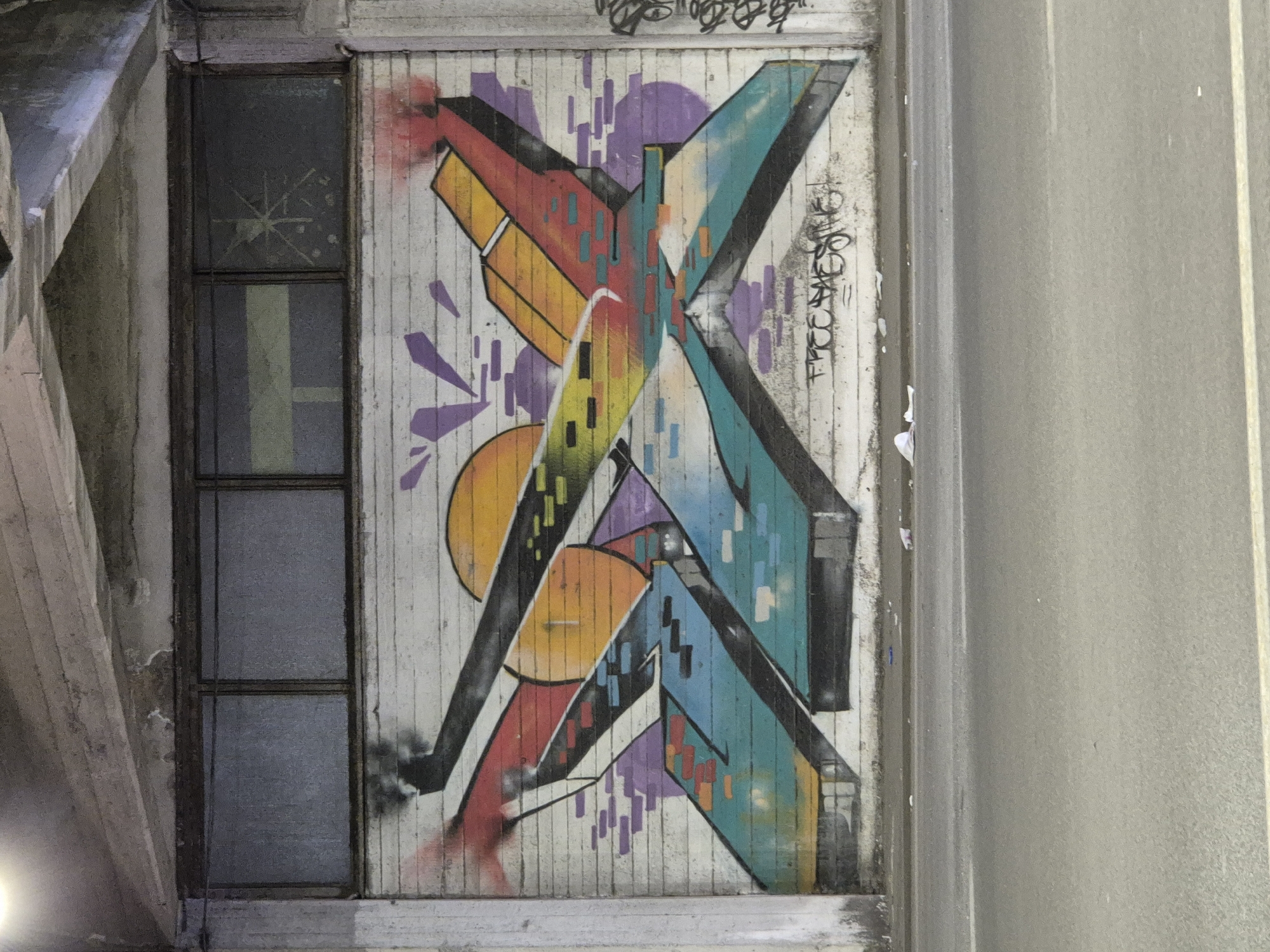 The image shows a section of a concrete underpass or bridge in Taranto, Italy, featuring vibrant graffiti art. The graffiti is a stylized representation of a large, angular "X" shape, rendered in a variety of bright colors including teal, yellow, orange, red, and purple. The artwork is painted on a horizontally striped white wall. Above the graffiti, there are several large, dark windows with visible reflections. The ceiling of the underpass, made of rough concrete, is visible at the top of the frame. The ground in front of the wall is a paved road, with some litter scattered near the base of the wall. To the right of the main graffiti, there is another section of graffiti in black marker, partially obscured. The lighting suggests it is nighttime or dusk. There are no people visible in the image, and the scene appears static.