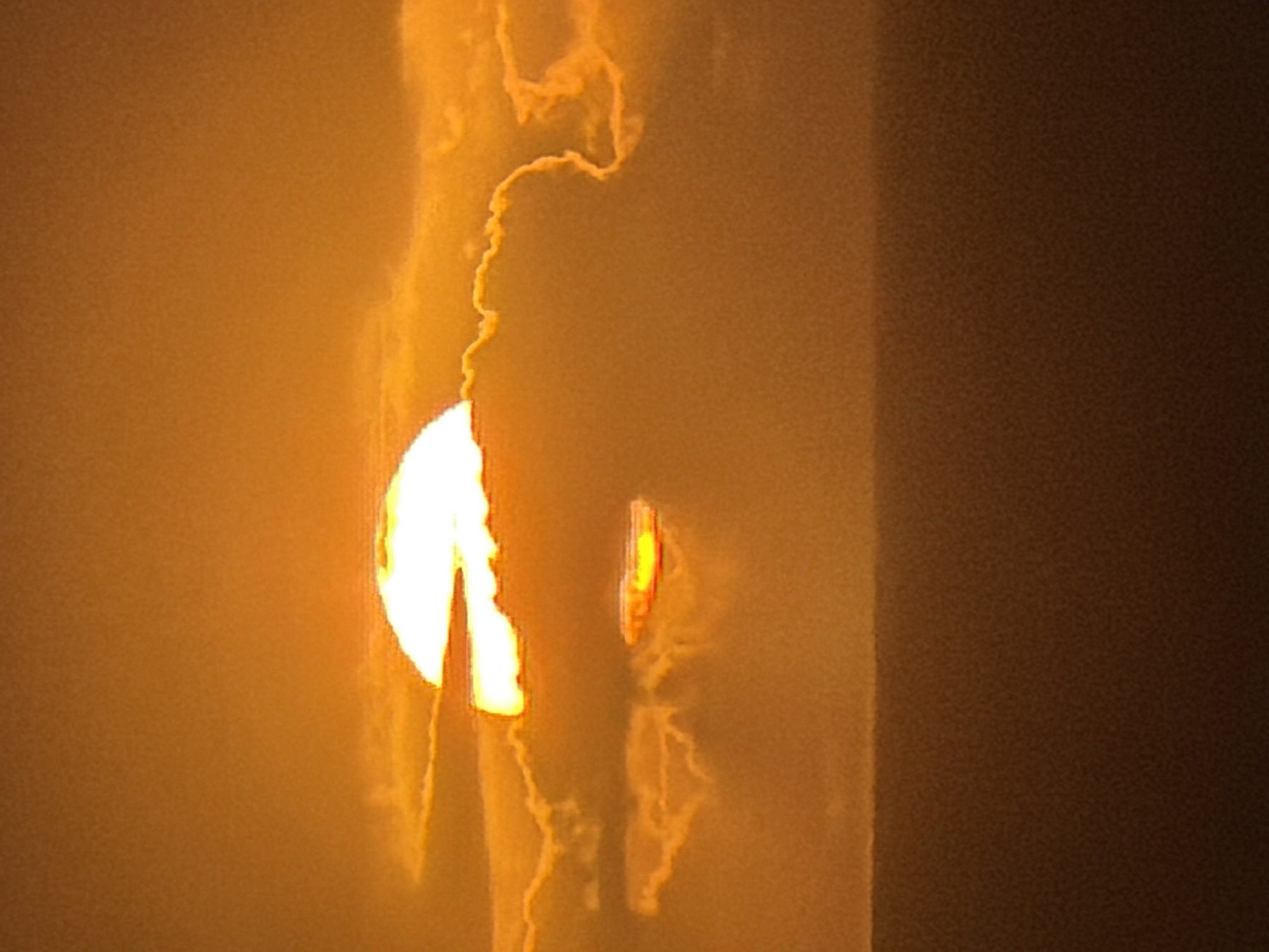 This image captures a dramatically lit landscape, likely at sunrise or sunset, in Cape Town, South Africa. The primary subject is an intensely bright, partially obscured circular light source, strongly resembling the sun, positioned on the left side of the frame. Its lower portion is hidden, suggesting it is either rising, setting, or partially covered by an unseen element.
Surrounding and extending upwards from this bright orb are abstract, wispy, and somewhat jagged formations of light orange and yellow. These formations appear to be clouds, heavy atmospheric haze, or smoke plumes, creating an ethereal and dynamic pattern against the warm-toned sky. To the right of the main light source, and slightly below its center, a smaller, elongated, and very bright light is visible. This could be a reflection, a secondary light source, or a distortion of the main light through the atmospheric conditions.
The rightmost section of the image is dominated by a dark, solid, vertical mass, appearing as a large structure, mountain, or landmass that partially obscures the scene and contrasts sharply with the vibrant light. The entire scene is enveloped in a thick, warm-toned haze, creating a monochromatic palette of bright yellows, oranges, and deep browns. This heavy atmospheric condition lends a powerful and mysterious ambiance to the setting. No people, discernible objects other than the light sources and atmospheric elements, or text are visible. The time of day is clearly dawn or dusk, and the weather is characterized by significant haze or smoke.