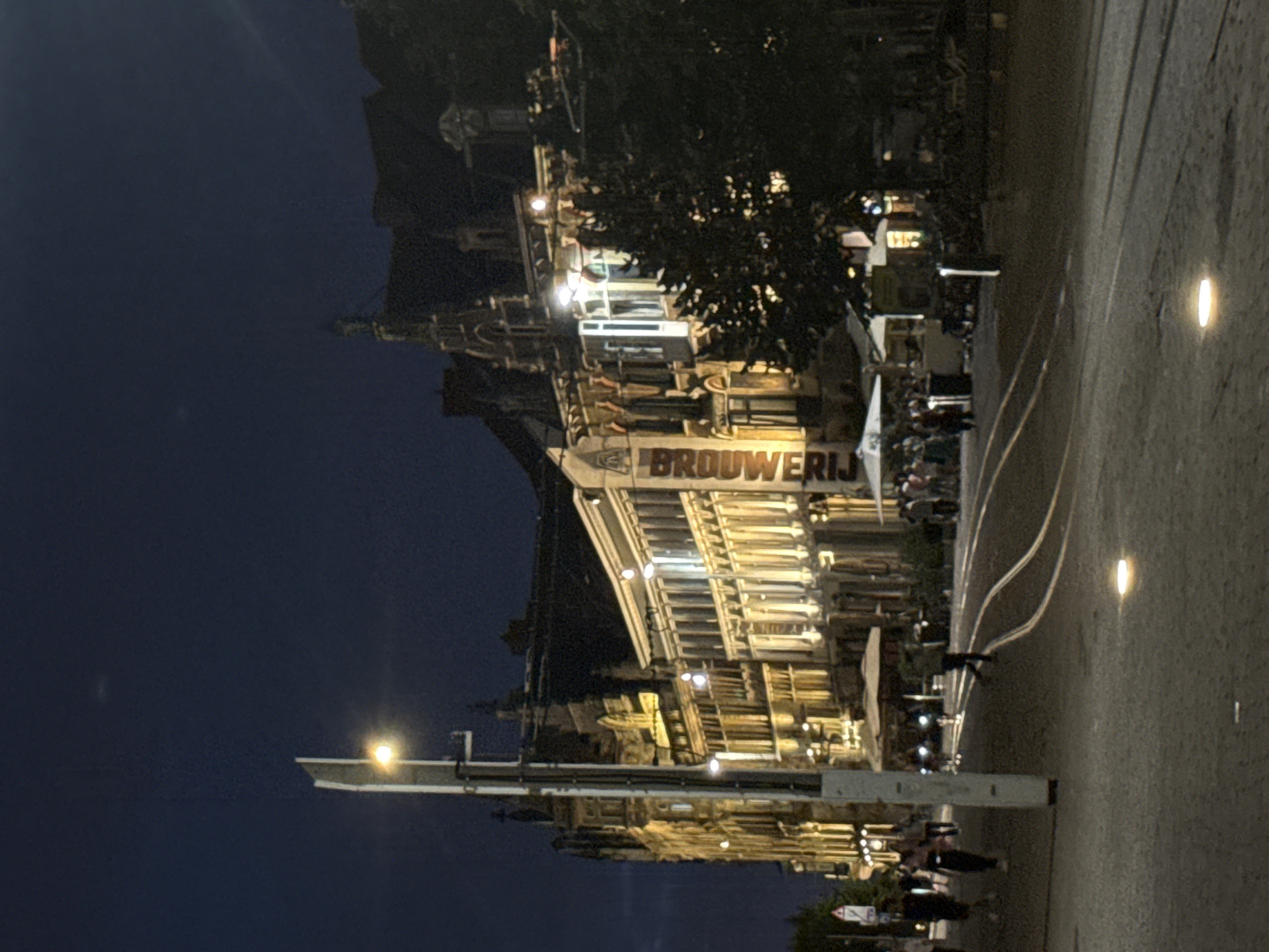 The image captures a nighttime view of a city street in Ghent, Belgium. In the foreground, a paved street with tram tracks is visible, illuminated by embedded lights in the pavement and a tall streetlamp to the left. Several people are walking or standing on the sidewalk. Behind them, a row of historic buildings with ornate facades and illuminated windows line the street. Some of these buildings appear to be restaurants or cafes with outdoor seating areas covered by umbrellas. The sky above is dark, indicating it is nighttime. The overall scene conveys a lively urban atmosphere at night.