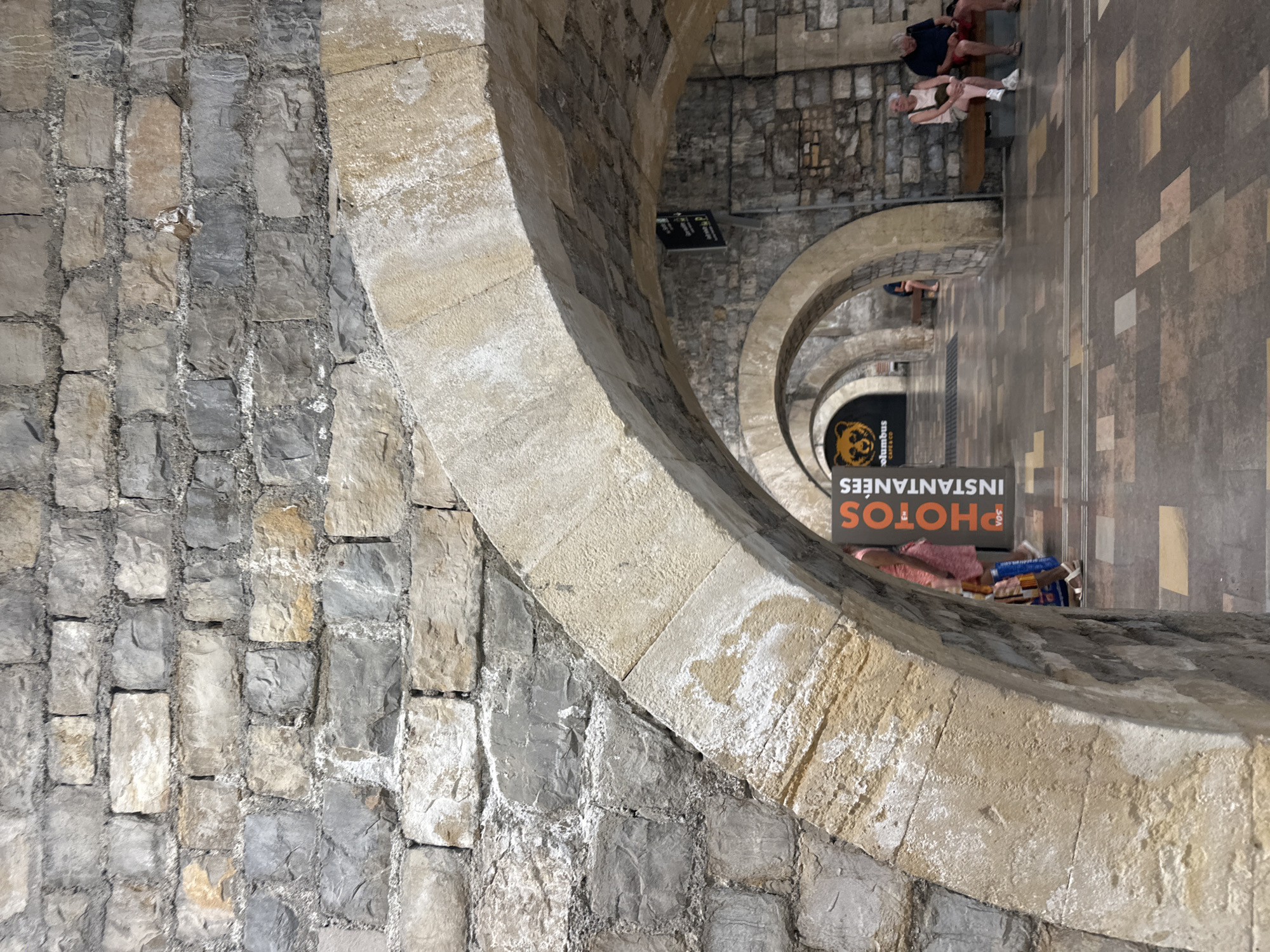 The image shows a perspective view looking through a series of arched stone structures. The foreground is dominated by a textured stone wall on the left and a smooth, lighter stone arch on the right. Beyond the foreground arches, more arches recede into the distance, creating a sense of depth.
In the background, along a tiled walkway, several people are visible. Some are sitting on a bench, appearing relaxed, while others are standing or walking. The scene suggests a public space, possibly a passageway or a covered walkway in an urban environment. The architecture, with its stone arches and tiling, hints at historical significance.
A prominent sign is visible hanging from one of the arches, featuring the words "PHOTOS INSTANTANEES" in orange lettering against a dark background. A stylized image of a bear's head is also part of the sign. This indicates a service related to instant photography is available in the vicinity.
The lighting suggests it is daytime, with light filtering through the arches. The overall impression is one of a well-trodden path, where people are going about their day, perhaps visiting or passing through a historic area of Nîmes, France. The stone textures and the repeating arch motif create a strong visual rhythm.