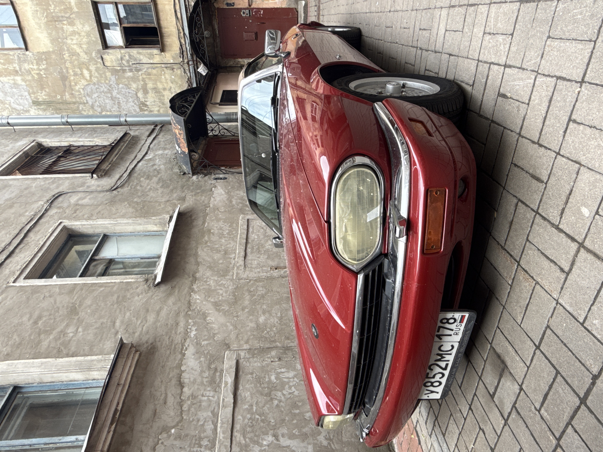 A red Jaguar XJS convertible is parked on a cobblestone street in a courtyard in Saint Petersburg, Russia. The car has a white license plate with Cyrillic characters and numbers. The building behind the car is old and weathered, with peeling paint and barred windows. There are two dark red doors on the building, one with an ornate metal canopy above it. The sky is overcast.
