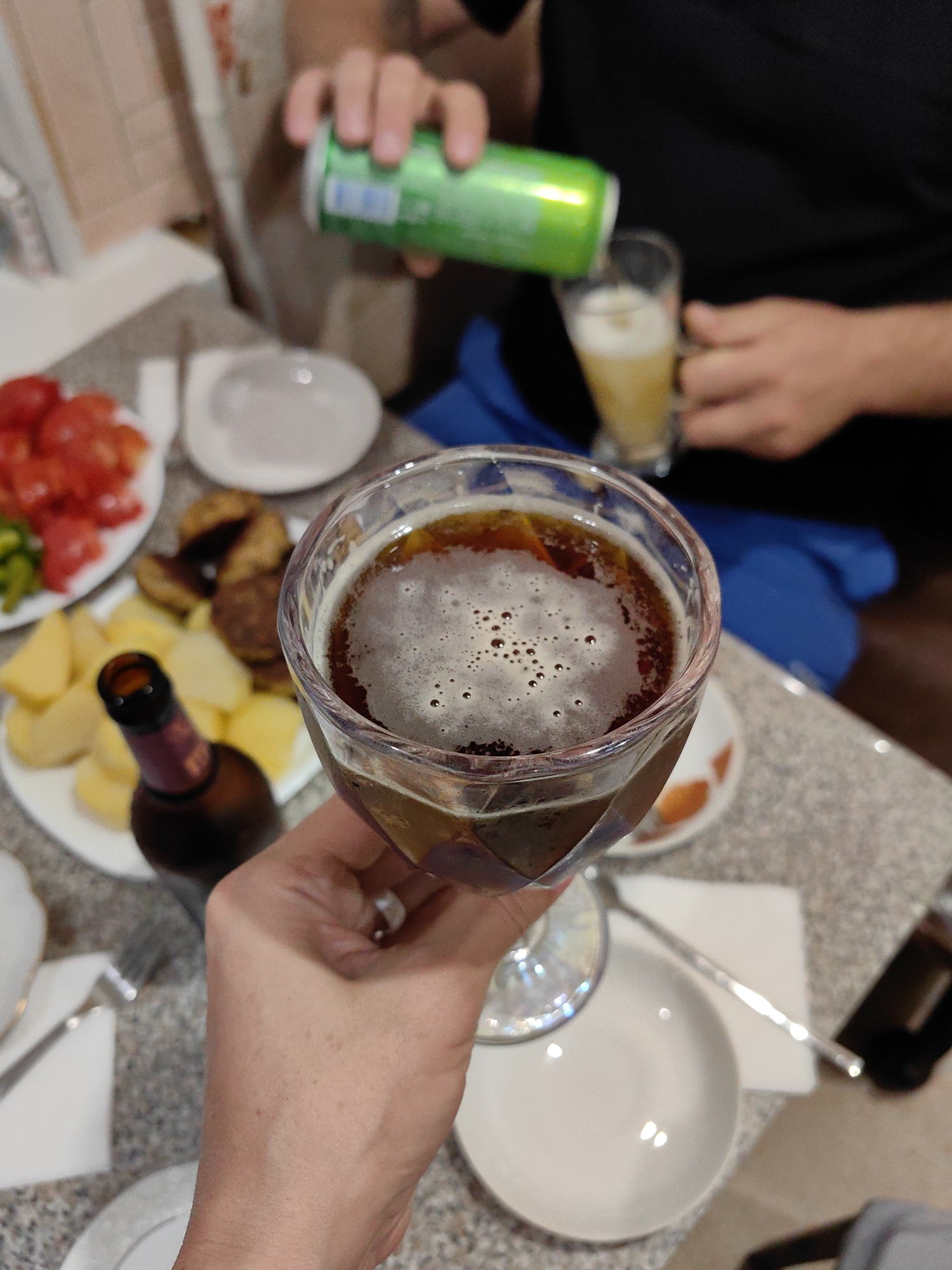 In this close-up, medium-shot photograph, a person's hand holds a stemmed glass filled with a dark liquid topped with a frothy head of foam. The glass is tilted, with the foam creating a dynamic pattern against the dark liquid. In the background, other elements of a meal are visible: a plate of sliced tomatoes, a plate with what appears to be cut potatoes and some form of meat patties, and a brown glass bottle. Another hand is seen in the upper background pouring from a green can into a tall glass of beer, indicating a social gathering with drinks. The setting is indoors, likely a dining area, with a patterned countertop and glimpses of other tableware. The image seems to be taken in Myadzyel, Belarus, suggesting a domestic or restaurant setting in that location.