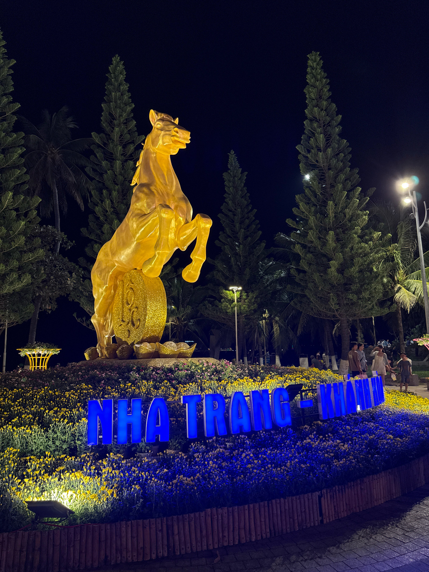 At night, a golden statue of a horse rearing on a large, ornate, circular base is illuminated in the foreground. The horse is positioned in front of several tall, dark green pine trees and palm trees. In front of the horse statue, a bed of yellow and blue flowers is arranged, with the words "NHA TRANG - KHANH HOA" spelled out in large, bright blue neon letters. Several people are visible in the background, walking and standing in a dimly lit area with streetlights. The scene appears to be a public space, possibly a park or a promenade, in Nha Trang, Vietnam.