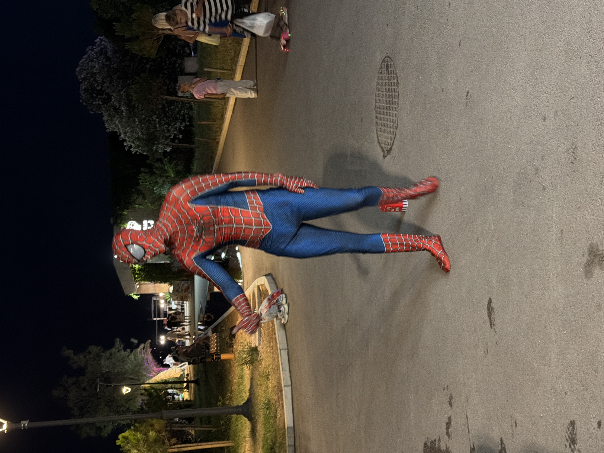 In an outdoor setting at night, a person dressed in a full Spider-Man costume stands on an asphalt pathway. The costume is predominantly red and blue with black web detailing on the red portions. The individual is captured mid-motion, with their right arm extended slightly forward and their left hand by their side. In the background, several other individuals are visible, some walking and others standing. One person, possibly a child, is wearing a pink shirt and white pants. Another group includes adults and a child. The environment appears to be a public area with trees, lighting fixtures, and a building in the distance. The ground is paved, and a manhole cover is visible in the foreground.