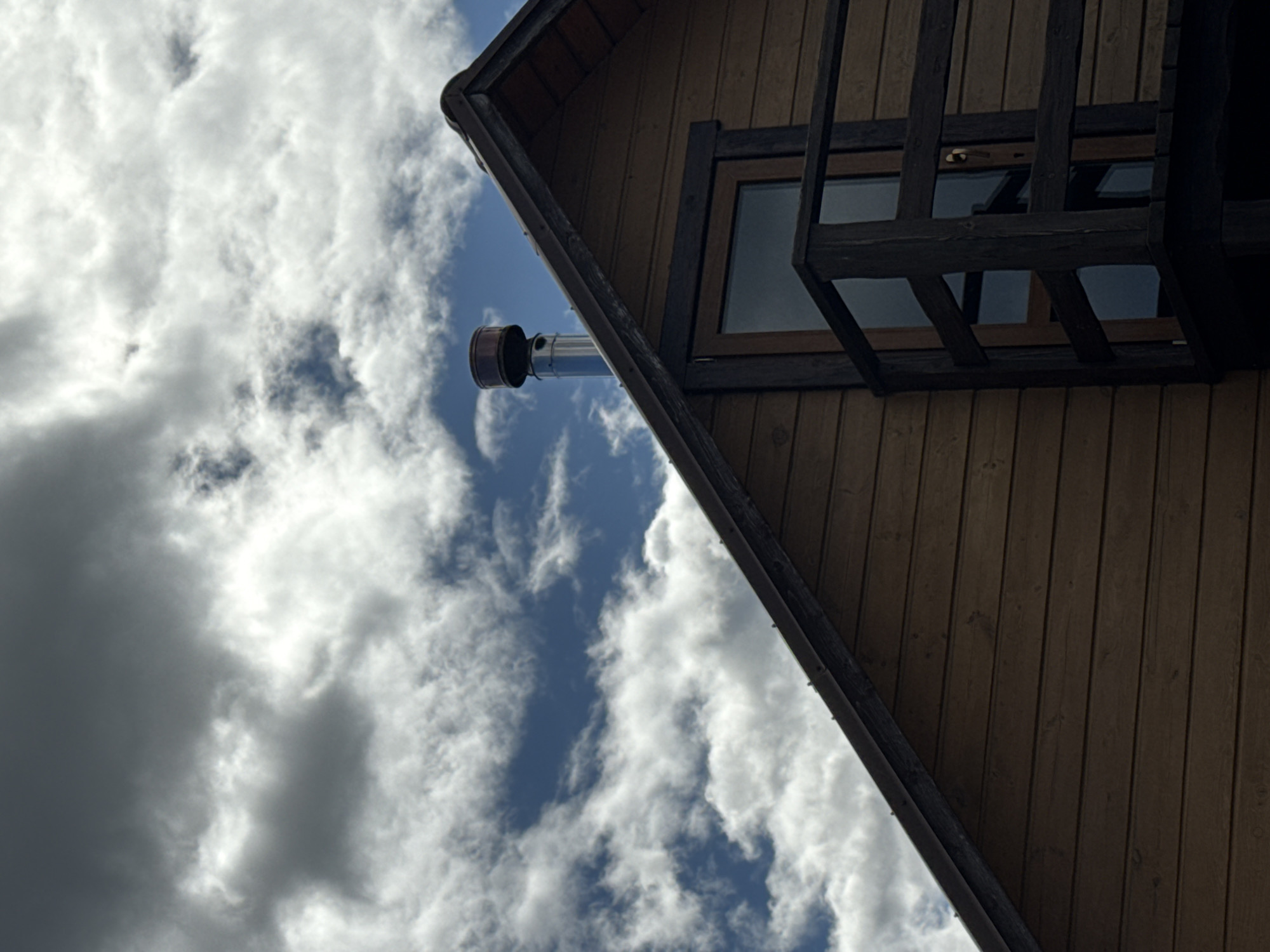 A low-angle shot depicts a brown wooden building with a chimney against a sky filled with white and gray clouds interspersed with patches of blue sky. A window with a dark frame and a dark wooden balcony can be seen on the side of the building. The building's roofline is angled from the lower left to the upper right of the frame. The photograph appears to have been taken outdoors. While the location is identified as Chaikovsky, Russia, there is no visible information in the image that directly confirms this.