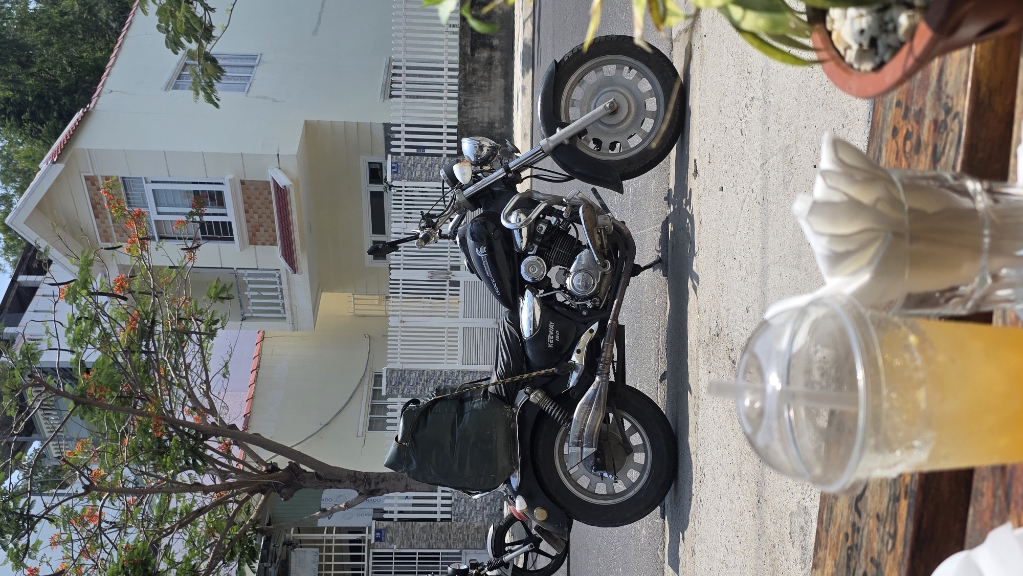 The image shows a black Keeway 150 motorcycle parked on the street in Bac Nha Trang Ward, Vietnam. In the background is a two-story house with a light yellow facade and a white picket fence. A tree with orange flowers stands near the motorcycle. On a table in the foreground, likely part of an outdoor seating area, are a plastic cup filled with a yellow beverage and a glass containing white tissues.