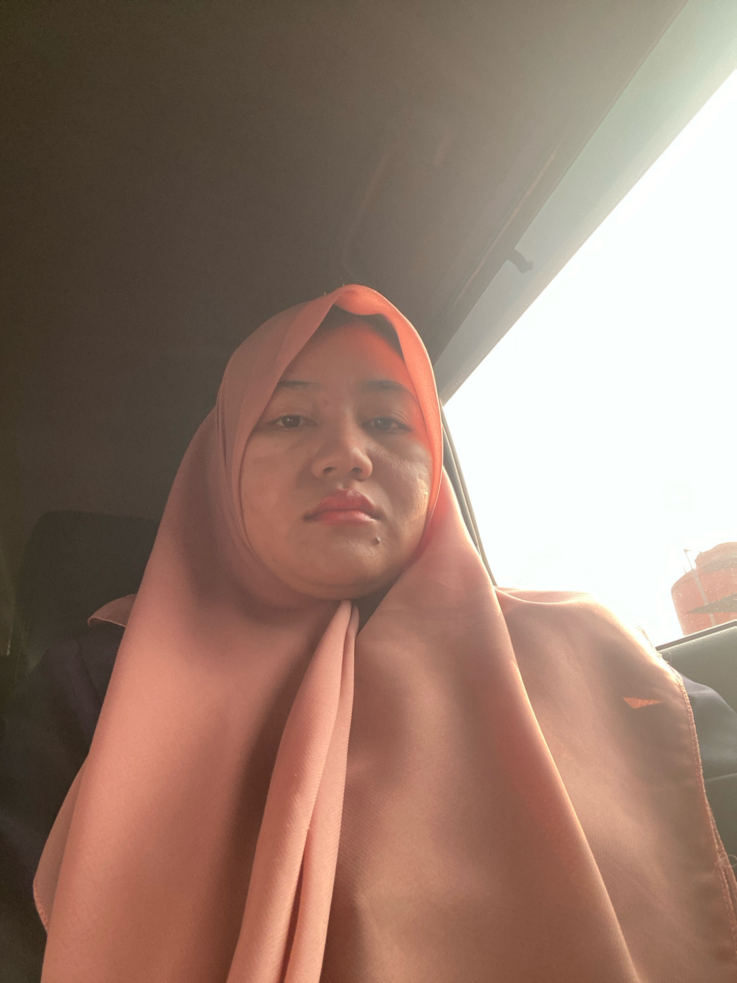 A medium shot captures a woman wearing a light pink hijab and dark clothing, looking directly at the camera. She is seated in the interior of a vehicle, with the dark interior visible on the left and a brightly lit window on the right. The window on the right shows a blurry view of an orange structure, possibly a building or a water tower, against a bright sky. The overall lighting suggests it is daytime, with strong light coming from the right side of the frame, creating some glare. The woman's face is illuminated from this light source. The background is dark and indistinct, typical of a car's interior. The image appears to have been taken from a selfie perspective. The location in Banjarbaru, Indonesia, is indicated by contextual information.