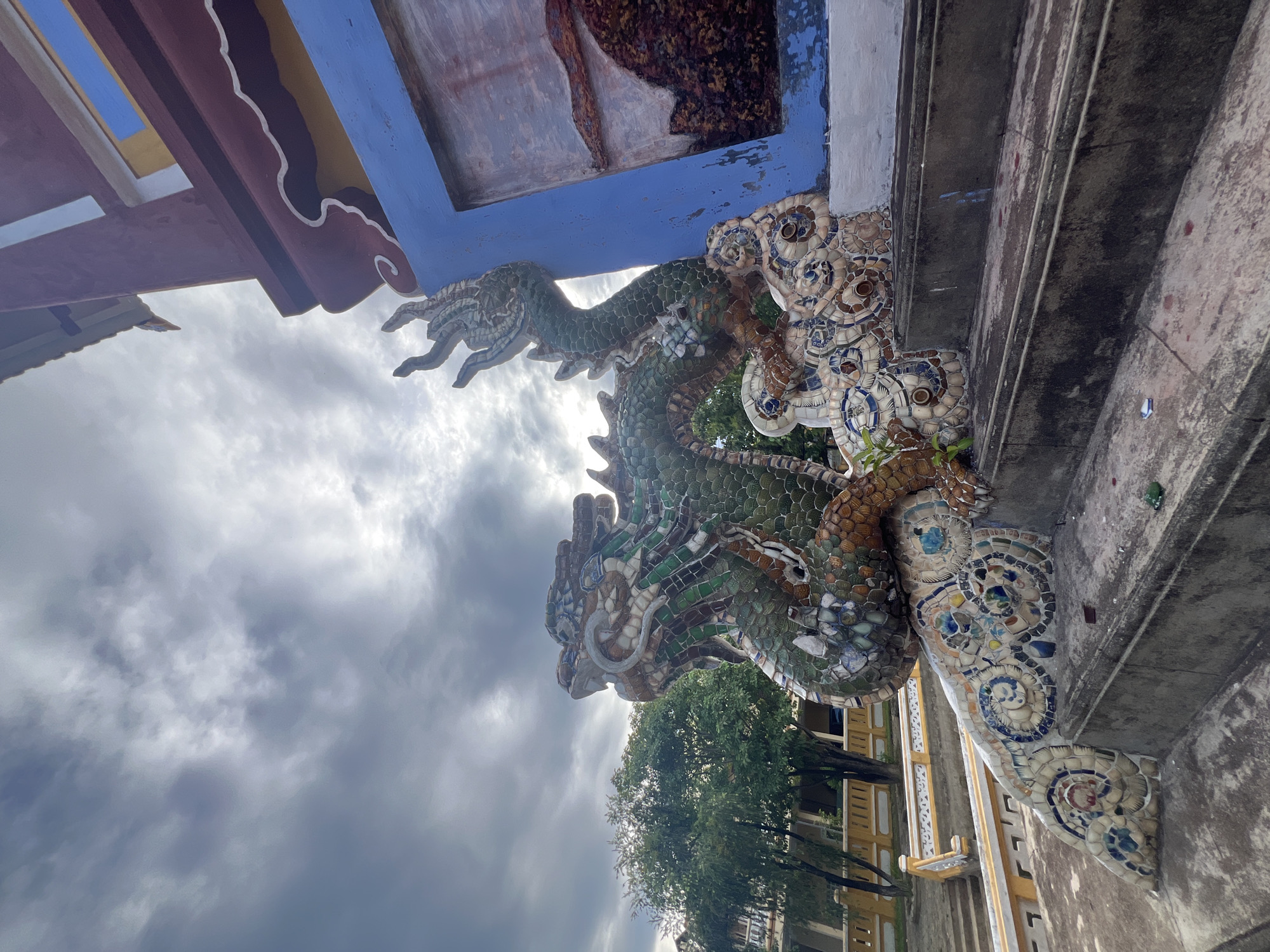 This image depicts a large, ornate mosaic dragon sculpture adorning the steps of a building. The dragon is crafted from numerous small, colorful tiles, predominantly in shades of green, brown, and white, with accents of blue and yellow. Its body is coiled around the steps, with its head raised and facing to the left. The dragon's scales and features are intricately detailed.
The building behind the sculpture features vibrant blue and red painted sections, typical of traditional Vietnamese architecture. The roofline is visible at the top, with decorative eaves. To the left of the dragon, a yellow railing and more steps are visible, leading towards a courtyard area with trees and other structures in the background. The sky above is cloudy, with dramatic, grey clouds suggesting an overcast or impending weather change. The foreground shows the concrete steps themselves, with some debris scattered on them.
While the specific location is not explicitly stated in the image, the architectural style, the presence of a mosaic dragon, and the overall ambiance are highly characteristic of the many temples and pagodas found in the ancient town of Hoi An, Vietnam. These detailed dragon sculptures are a common decorative element in such religious and historical sites within Hoi An.