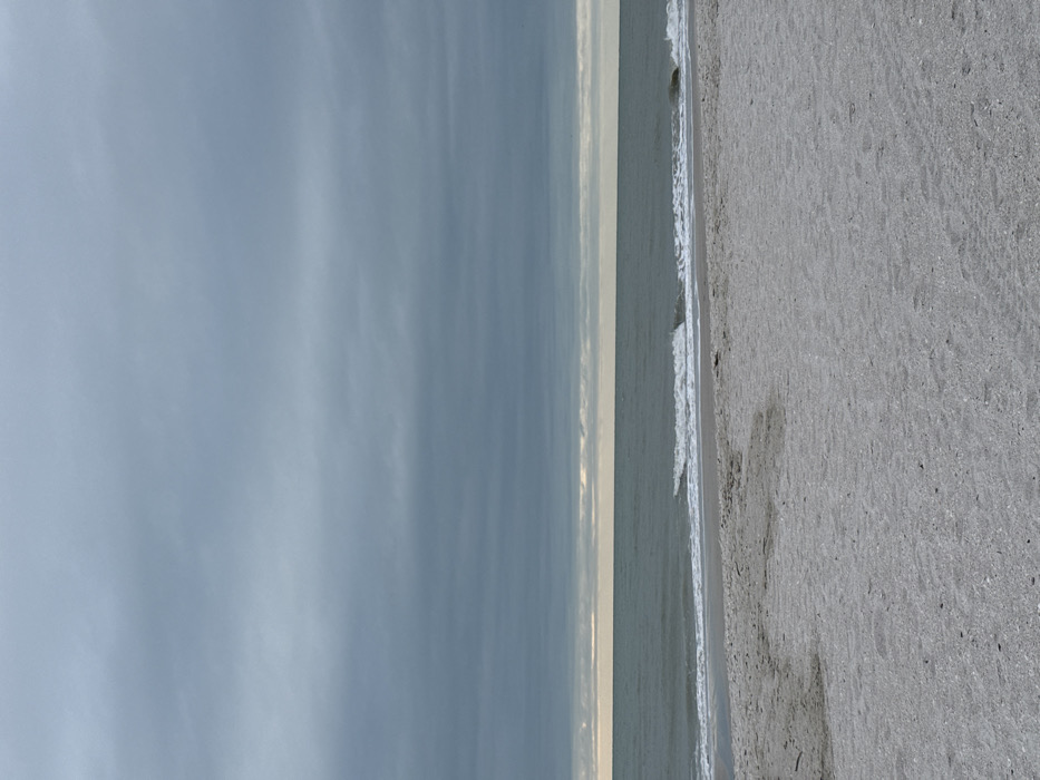 The image displays an uninhabited beach scene in Naples, United States of America, under an overcast sky. The upper two-fifths of the frame is occupied by a dense, uniform layer of dark grey clouds. A narrow horizontal band of pale yellow-grey light is visible at the horizon, situated between the cloud cover and the ocean. The middle section of the image features the ocean, characterized by dark grey-green water, with multiple small waves capped by white foam approaching the shore. The lower two-fifths of the frame shows an expansive area of light grey-white sand beach. No individuals, animals, or man-made objects are visible, and no specific actions or interactions are depicted within the scene.