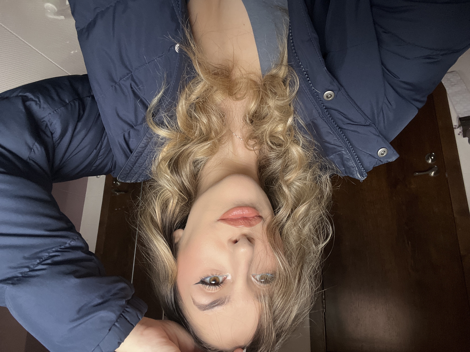 A person with blonde curly hair, wearing a blue padded jacket over a grey top, is photographed from an upside-down perspective. The background reveals a dark wooden door with a silver handle, as well as a white-tiled wall. The location is not discernible from the image.