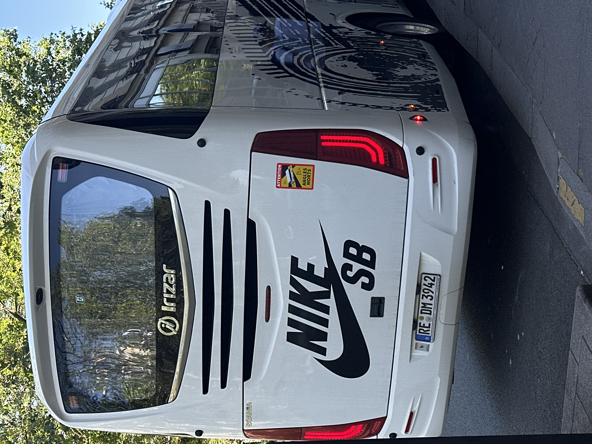 A brightly-lit, daytime photograph taken at street level features the rear of a white Irizar bus parked along a street. The bus displays a large Nike SB logo, along with the license plate "RE DM 3942" and a sticker indicating blind-spot awareness in French ("Angles Morts"). Reflections in the bus window reveal a street scene with trees and buildings. The location is strongly suggested to be Paris, France, based on the style of the building architecture. The bus is positioned parallel to the curb with the rear facing the camera, slightly angled toward the street.