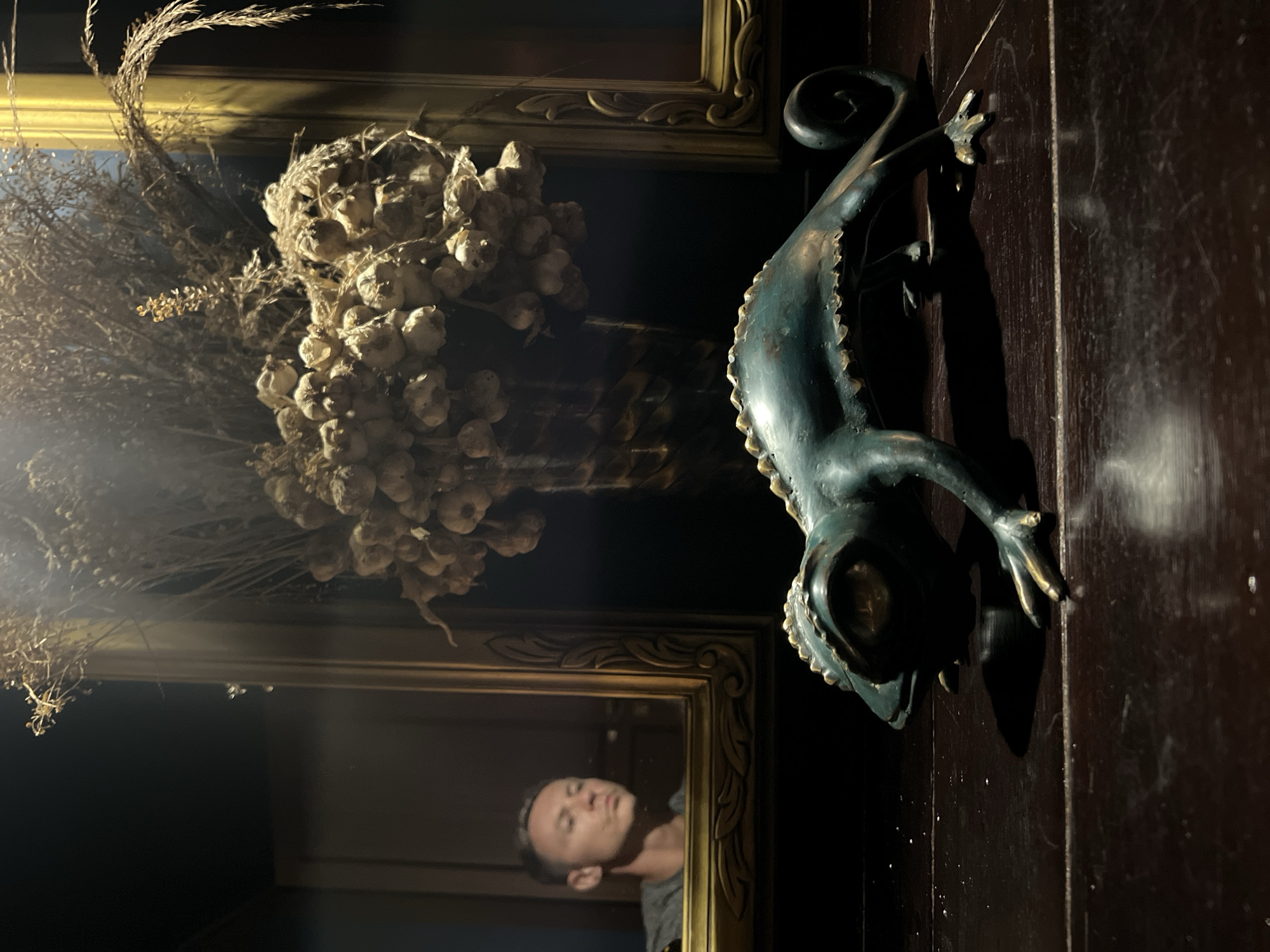 A still-life image taken indoors in Ubud District, Indonesia, features a bronze-colored sculpture of a gecko, placed on a dark wooden surface. To the left of the gecko is a framed arrangement containing dried plant matter, including garlic bulbs and sprigs. The lower frame reflects the image of a man. The lighting is dim, with a bright area that creates a vignette effect.