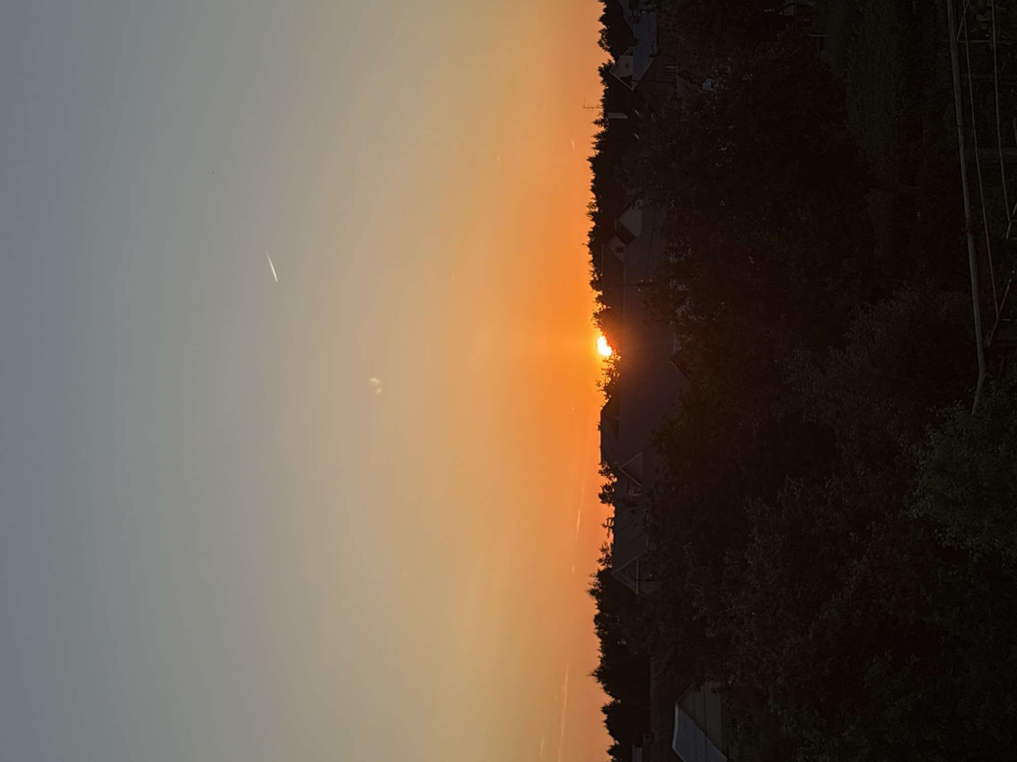 The image depicts a sunset over a semi-rural area, likely a dacha community or a small settlement on the outskirts of a city, possibly within or near Прикамский, Russia. The sky transitions from a pale blue at the top to a vibrant orange glow near the horizon, characteristic of a sunset. A faint contrail from an aircraft is visible in the upper portion of the sky.
In the mid-ground, the silhouettes of numerous trees form a dark, dense line against the bright sky. Among the trees, the rooftops of several houses and buildings are visible, appearing as dark shapes with some roof details discernible. The architecture suggests residential structures, possibly with pitched roofs.
The foreground is dominated by the dark silhouettes of lush foliage, likely trees and bushes, obscuring the ground level. A partial view of a railing or fence structure can be seen at the very bottom of the frame. The overall lighting is dim due to the sunset, with the brightest illumination concentrated around the horizon where the sun is setting. There are no visible people or specific actions taking place in the scene.