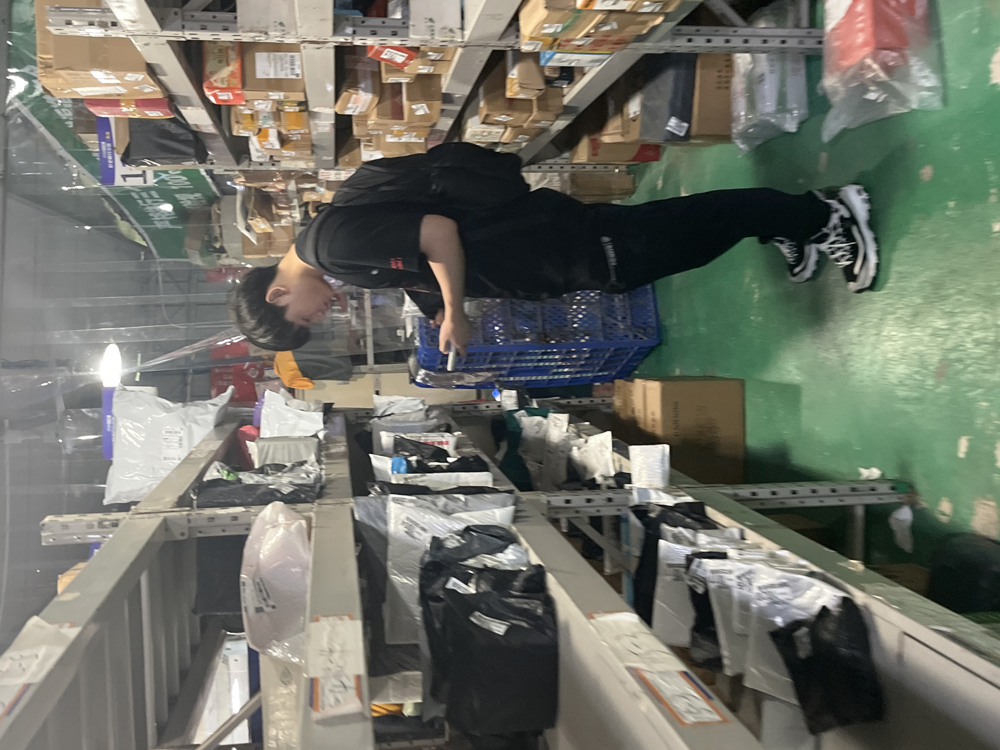 A person is standing in a warehouse-like environment filled with shelves loaded with boxes and packages. The individual, dressed in black clothing, is holding a mobile phone. The shelving units stretch into the distance, packed with items of various sizes and shapes. The floor is green. Based on the presence of Chinese text on a sign in the upper left corner of the frame (严打诈骗100), and a sign referencing Jiangjin in Chinese, this scene likely occurs in Jiangjin, China.