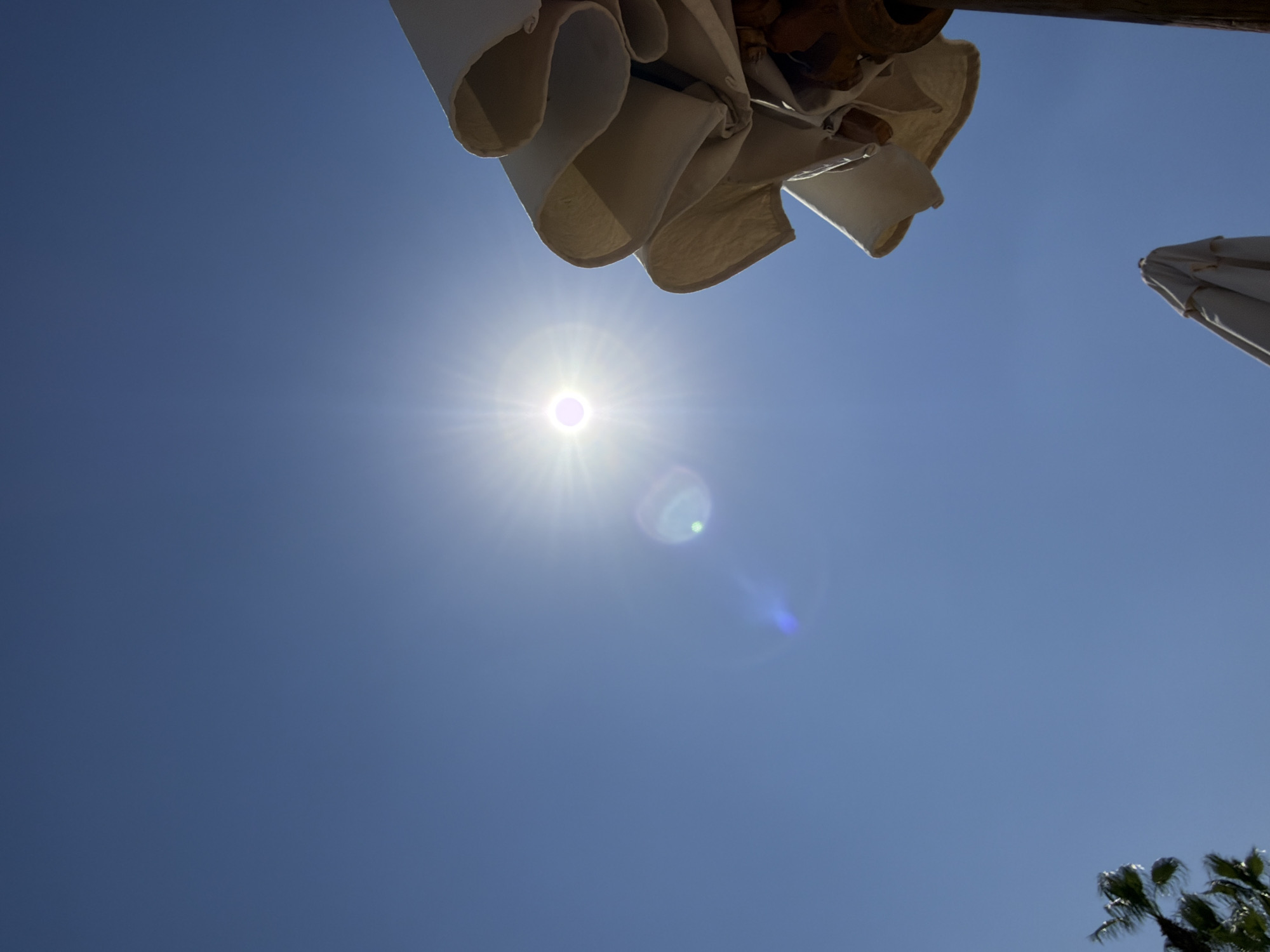 The image depicts a bright, sunny day with a clear blue sky. The sun is prominently positioned in the upper center of the frame, emitting a strong glare and lens flare. In the foreground, partially obscuring the sky on the right side, are the folded canopies of two beach umbrellas. The umbrella in the upper right is partially open, revealing its textured fabric and wooden frame. The second umbrella, closer to the bottom center, is fully closed. In the lower left corner, the fronds of a palm tree are visible, suggesting an outdoor, possibly tropical or subtropical, setting. The overall impression is of a summery day at a beach or resort. The image is consistent with the environment found in Sharm El Sheikh, Egypt, a popular tourist destination known for its sunny climate and beaches.