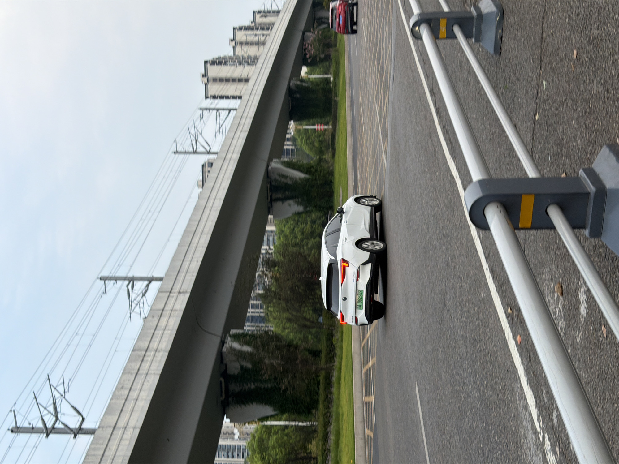 The image depicts an overpass with power lines and a road with two cars, one white and one red, traveling in the same direction. A white station wagon is in the foreground, on the right side of the road. It has its brake lights illuminated and a license plate reading "A DB2909". The red car is in the distance, also on the right side of the road. The road surface is asphalt, with visible lane markings and a pedestrian crossing marked with yellow lines. The overpass is a concrete structure with supporting pillars. In the background, several multi-story buildings are visible, as well as trees and vegetation. The sky is overcast. The scene appears to be located in Jiangjin, China, specifically on a road underneath a railway or highway overpass, given the presence of the power lines and the architecture of the surrounding buildings.