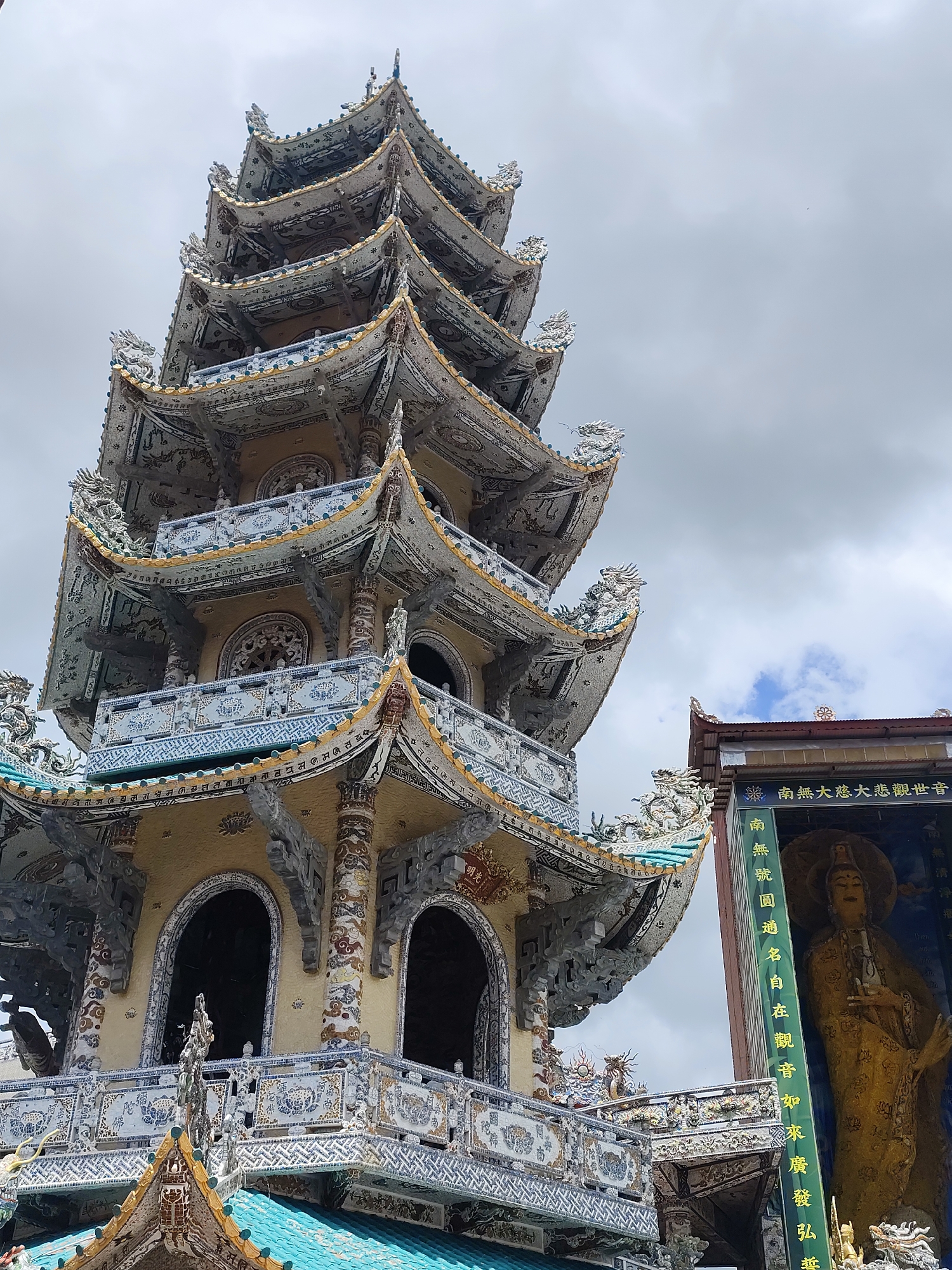 The image shows the Linh Phuoc Pagoda in Xuân Thành, Vietnam. The pagoda is a tall, multi-tiered structure with intricate mosaic decorations featuring dragons and other mythological creatures. The building is adorned with various colored tiles and ceramics, giving it a vibrant and ornate appearance. To the right of the pagoda, a large statue of a deity, likely Guanyin, is visible within an alcove. The statue is golden and depicted with a serene expression. The sky above is overcast with some patches of blue sky and clouds visible. The foreground features architectural details of the pagoda, including balconies and arched windows.
