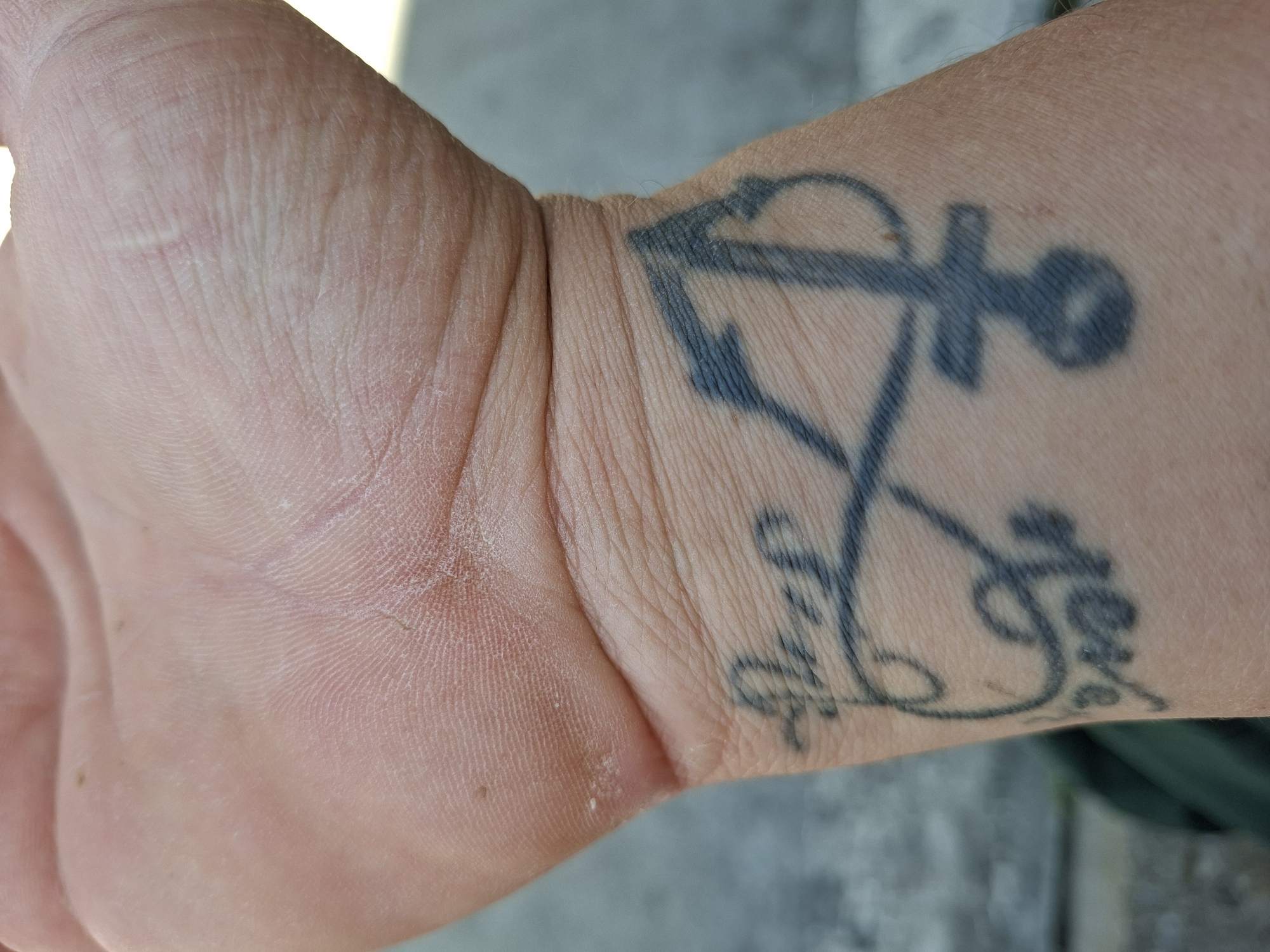 A close-up image depicts a person's wrist and hand. On the wrist, there is a tattoo featuring an anchor design intertwined with cursive lettering that spells "Faith" and "Love". The skin shows fine wrinkles and texture consistent with age. The background is out of focus, with hints of grey or light green. The image does not provide any information about the location in Plovdiv, Bulgaria.