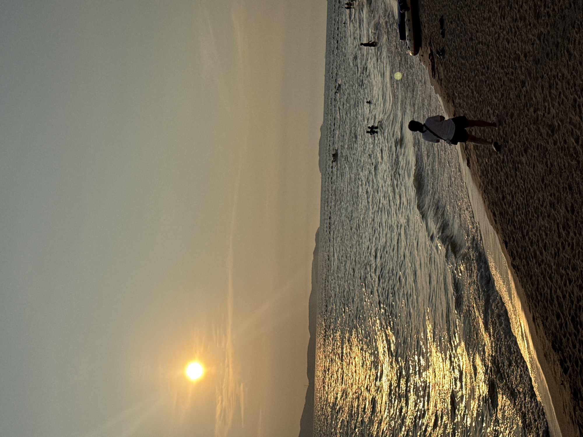 The image depicts a beach scene at sunset in Pattaya City, Thailand. The sun is low in the sky, casting a golden light across the water and the sand. There are several people in the water, and one person standing on the beach. The waves are small and gentle. The sky is hazy with a yellow and orange hue.