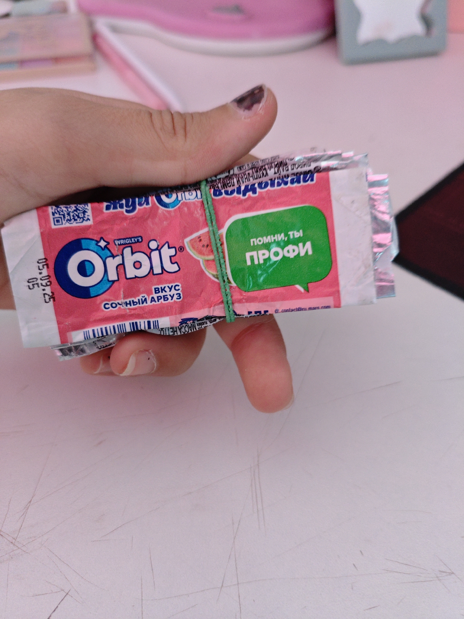 A hand is holding a pack of Orbit chewing gum, flavored with juicy watermelon. The gum pack is pink and features the Orbit logo, watermelon slices, and text in Russian that translates to "Remember, you're a pro." A green rubber band is wrapped around the gum pack and the hand. The background shows a pink surface, possibly a desk, with some blurry objects. The image does not provide any information about the city of Трудовское сельское поселение, Ukraine.