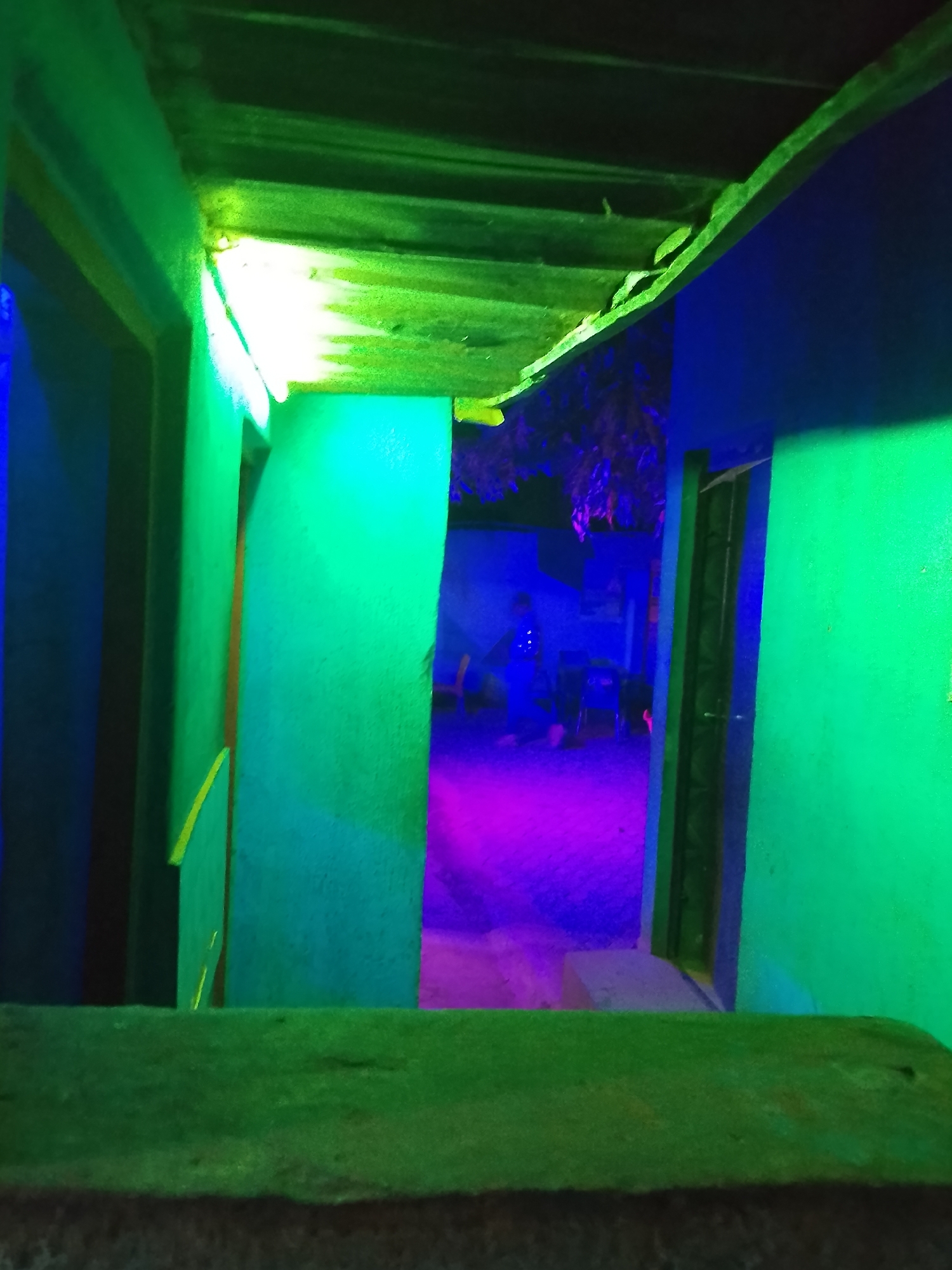 Captured from within a structure, the image provides a view of an outdoor space bathed in a vibrant and unnatural palette of blues, greens, and purples. The vantage point is within a building with walls painted in shades of green and blue, and a rough ceiling made of stacked beams. An entryway or opening in the building frames the outdoor scene. Outside, the ground appears to be illuminated with a strong purple or magenta light, suggesting an artificial source. There are blurry figures in the background, one possibly seated on a chair, and what appears to be foliage above, bathed in blue light. The overall atmosphere is surreal and artificially lit, devoid of natural color representation.
