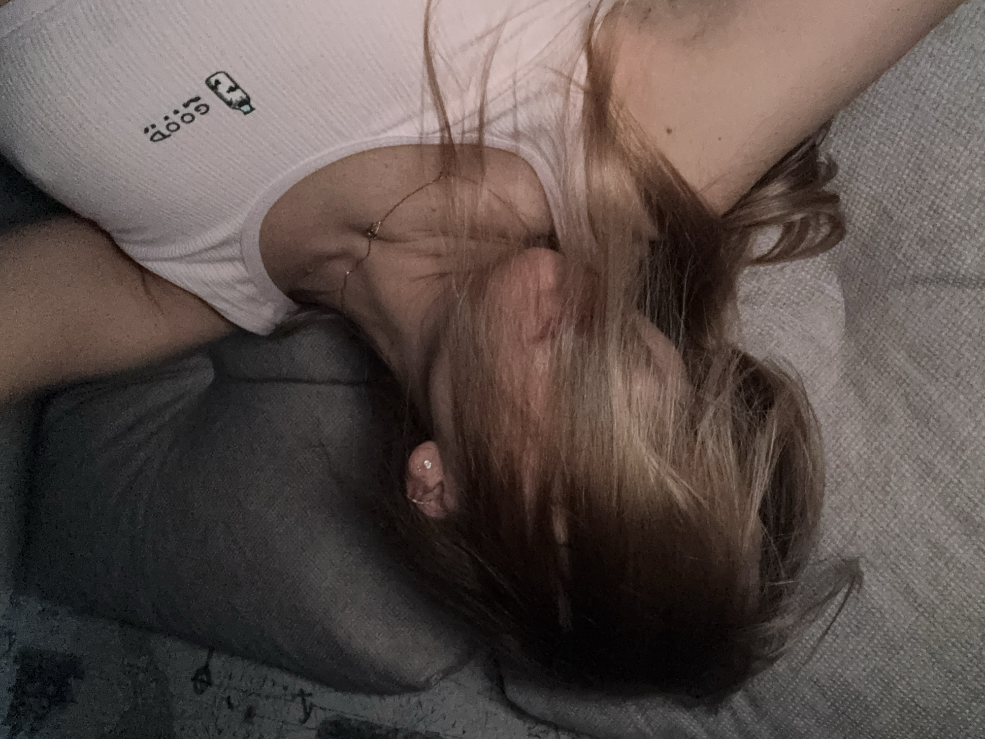 The image presents a close-up, high-angle view of a person, likely female, lying down indoors on what appears to be a gray sofa or bed, with their head resting on a gray pillow. The person's face is mostly obscured by long, light brown hair that spreads across their head and upper chest.
They are wearing a white, ribbed tank top or camisole. On the left shoulder area of the top, the word "GOOD" is visible in black text, styled with two small dots above each 'O', accompanied by a graphic of a stylized bottle or container. A delicate silver chain necklace with a small pendant is visible around the person's neck, and a small, sparkling silver-colored earring is present in their visible ear. The lighting in the scene is soft and ambient, suggesting an indoor setting with subdued illumination, potentially during the evening or in a dimly lit room. The overall atmosphere conveys a sense of calm and relaxation, with the person appearing to be resting. No specific visual cues within the image directly indicate the provided location context of Ust-Ilimsk, Russia.