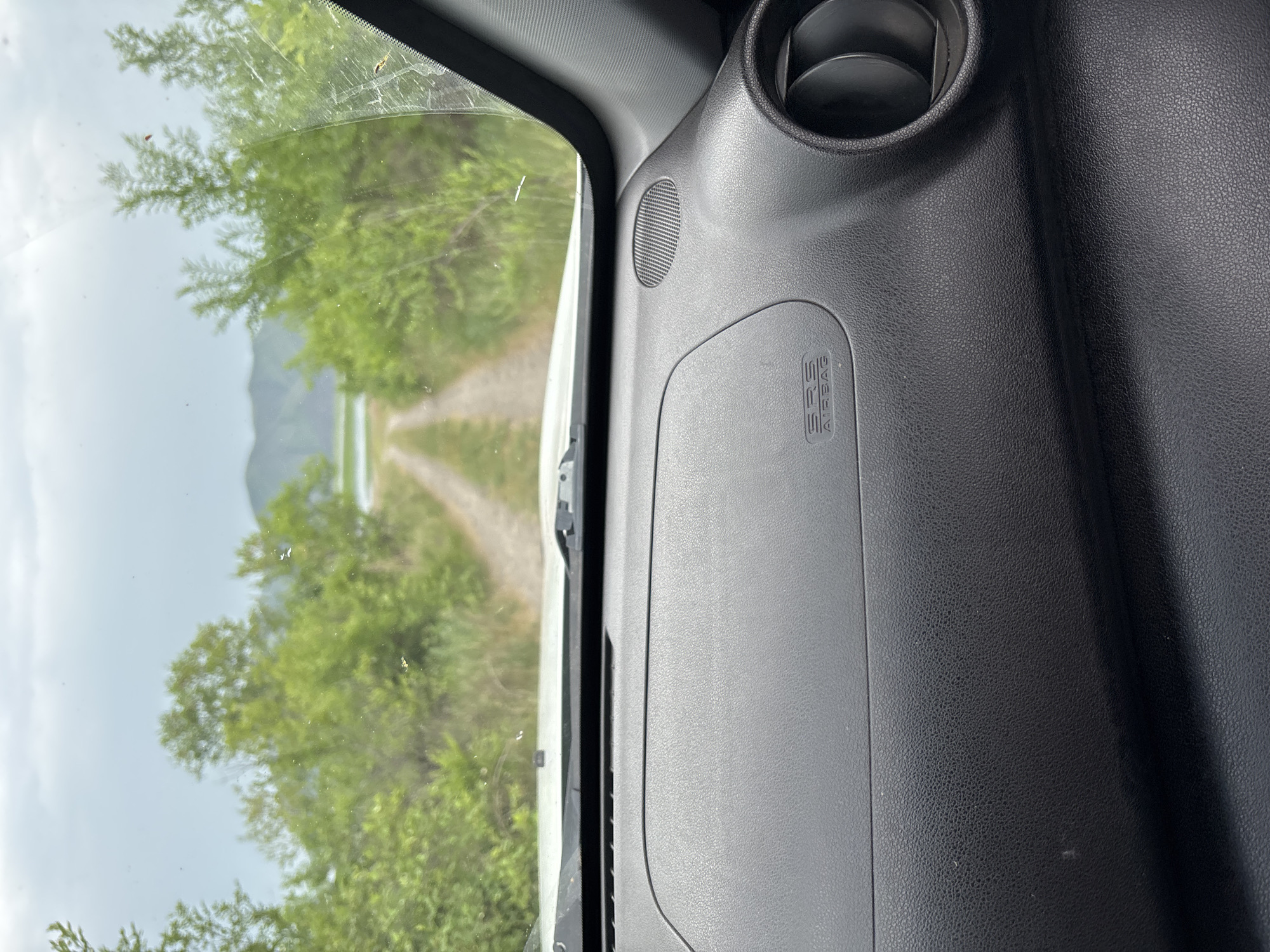 The image shows a view from inside a vehicle, looking out through the windshield. A dirt road leads into the distance, flanked by lush green trees and vegetation. In the background, a body of water is visible, with hills beyond it under a cloudy sky. The dashboard of the car is in the foreground, with an "SRS AIRBAG" label visible on the passenger side. The scene appears to be in a rural or natural setting, possibly on the outskirts of Chernogorsk, Russia, given the mention of the city. There are no people visible in the image, and the focus is on the natural landscape and the interior of the vehicle.