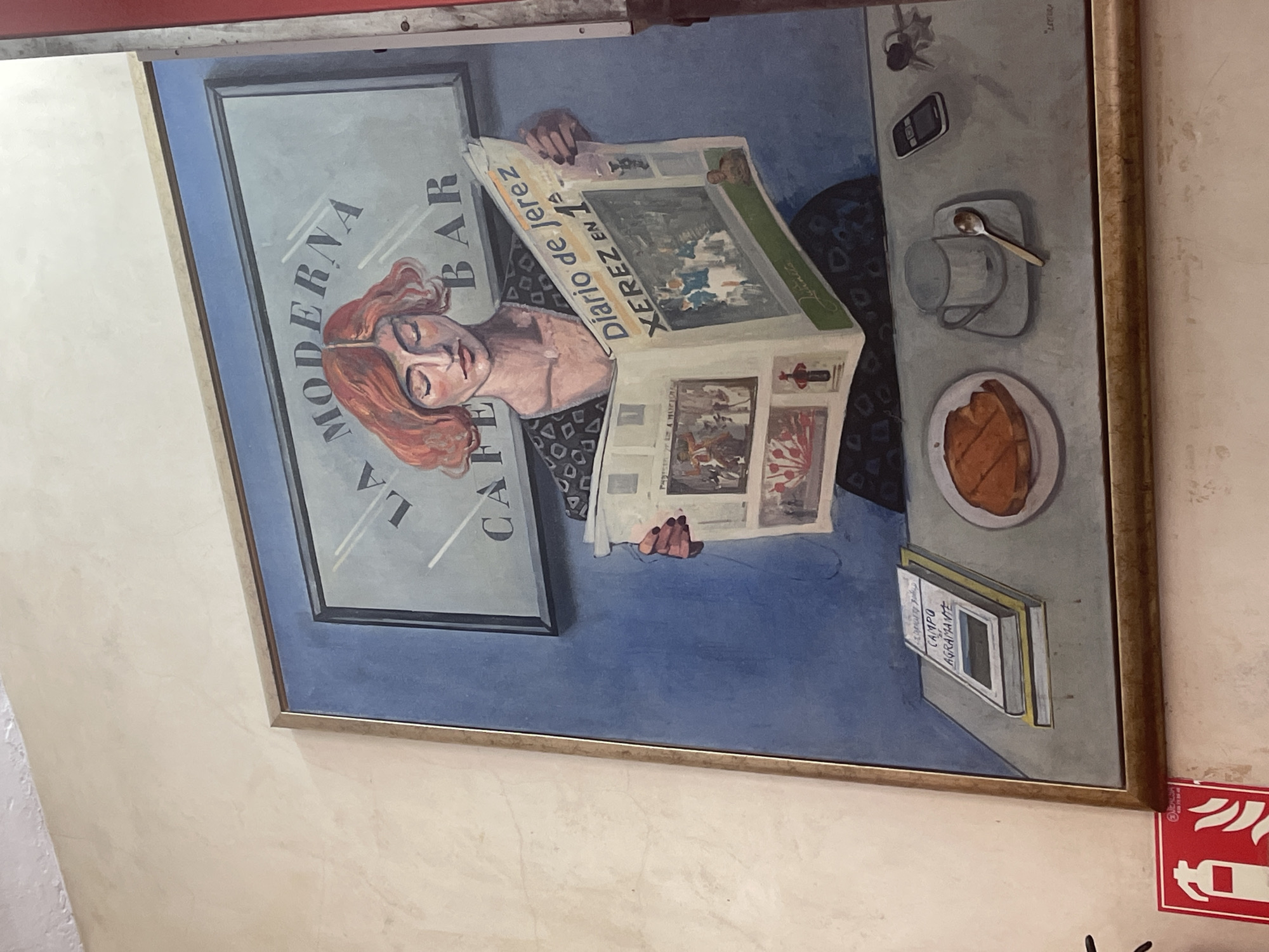 The image is a framed painting hanging on a wall. The painting features a woman with reddish-orange hair, seated at a table in what appears to be "La Moderna Cafe Bar," as indicated by the text in the painting. She is reading a newspaper titled "Diario de Jerez XEREZ EN 1ª." On the table, there's a cup with a spoon, a plate with what seems to be food, a cell phone, and a book titled "CAMPO AGRAMANTE." The background is a blue hue with the cafe bar signage. The frame of the painting is gold-colored. Adjacent to the lower right corner of the framed painting is a red fire extinguisher sign attached to the wall. The location mentioned is Jerez De La Frontera, Spain, based on the newspaper title.