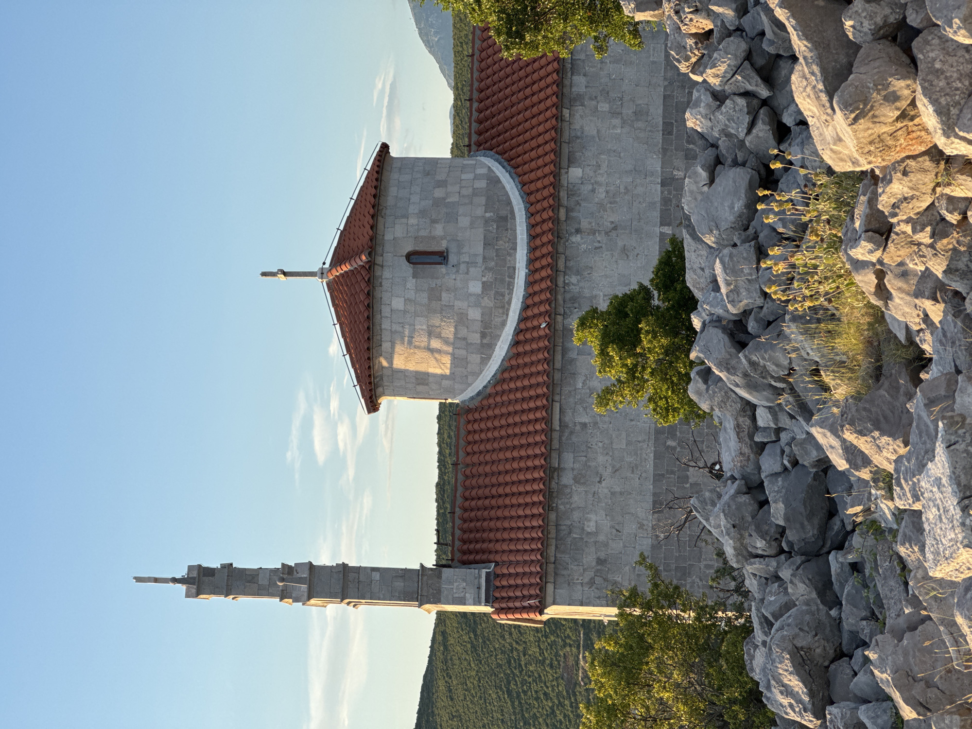 The image displays a stone church with a red-tiled roof, featuring a prominent bell tower or minaret on the left side and a dome-shaped section with a cross on top. The building is constructed with large, gray stone blocks, and a small, arched window is visible on the domed portion. The church is situated in a rocky, arid landscape with sparse green vegetation and trees. In the foreground, a dense pile of large, gray rocks creates a natural barrier. The background shows rolling, green hills under a clear blue sky with a few wispy clouds. The lighting suggests it is either early morning or late afternoon, with sunlight casting shadows on the building and highlighting the textures of the rocks and vegetation. The scene appears serene and isolated, with no people or animals visible. The location is identified as the city of Kovačko Polje, Montenegro.
