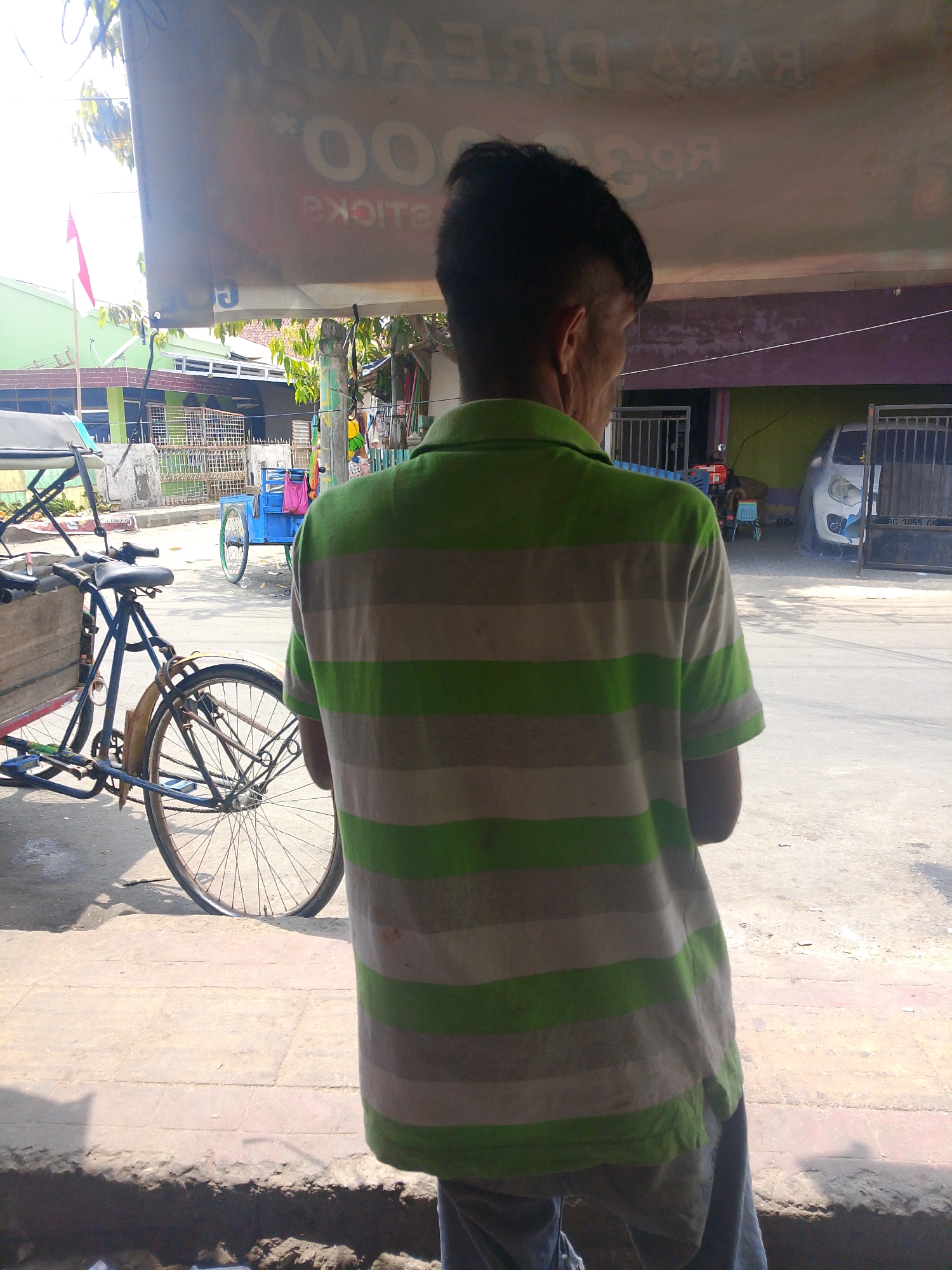 A person wearing a green and gray horizontally striped polo shirt stands with their back to the camera, facing a street scene in Palembang, Indonesia. They are positioned in the foreground, obscuring a part of the view. In the middle ground, a blue cart with a pink bag on it is visible on the street, and a bicycle rickshaw with a wooden cargo area and a large rear wheel is parked. Further in the background, a white car is parked in front of a building with a gated entrance. The environment appears to be a bright, sunny day. A large banner with Indonesian text is partially visible overhead, with the words "Y.M.A.S" and numbers possibly indicating prices. The banner also features a red and white Indonesian flag. The ground is paved with what appears to be concrete slabs. The overall scene suggests a commercial area or a street market.
