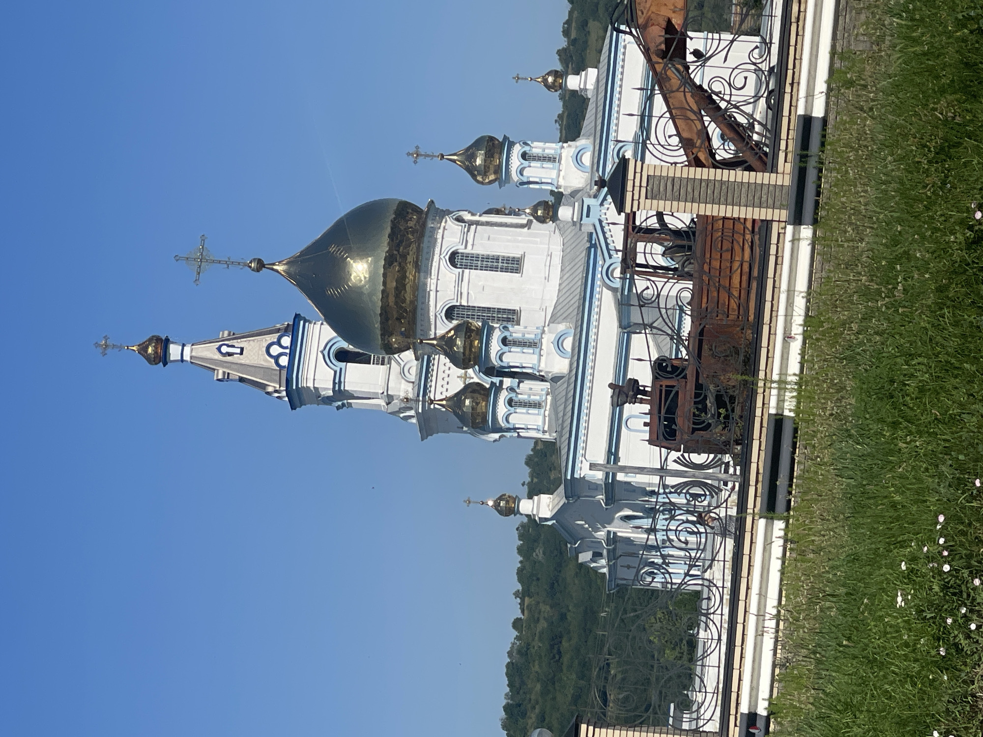This is a vertical, outdoor, medium shot of a white and blue Orthodox church with golden domes. The church has several onion domes, the largest of which is prominent in the center of the frame. The building is adorned with ornate blue trim and white window frames. Several crosses are visible atop the domes.
To the right of the church, a decorative black metal fence encloses an area with some weathered, rusted metal structures, possibly old decorative elements or machinery. Beyond the fence, a strip of green grass dotted with small white flowers is visible. The background is dominated by a clear, bright blue sky, with a distant line of lush green trees forming a gentle slope on the horizon. The sunlight suggests it is a bright, clear day, likely daytime given the clear sky and shadows. No people or text are visible in the image. The scene conveys a sense of peace and religious solemnity against a natural backdrop.