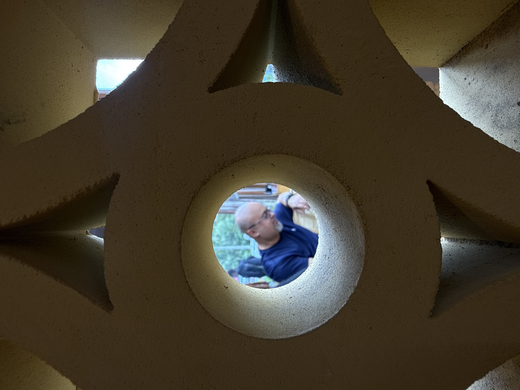 In Da Lat, Vietnam, a man with a shaved head and glasses peers through a circular opening in a textured, beige decorative concrete screen. He is wearing a dark blue t-shirt. His gaze is directed downwards, and his left hand rests on a surface. In the background, another person is visible, and beyond them, there is greenery and what appears to be a window. The scene is framed by the geometric patterns of the concrete screen, creating a layered effect.